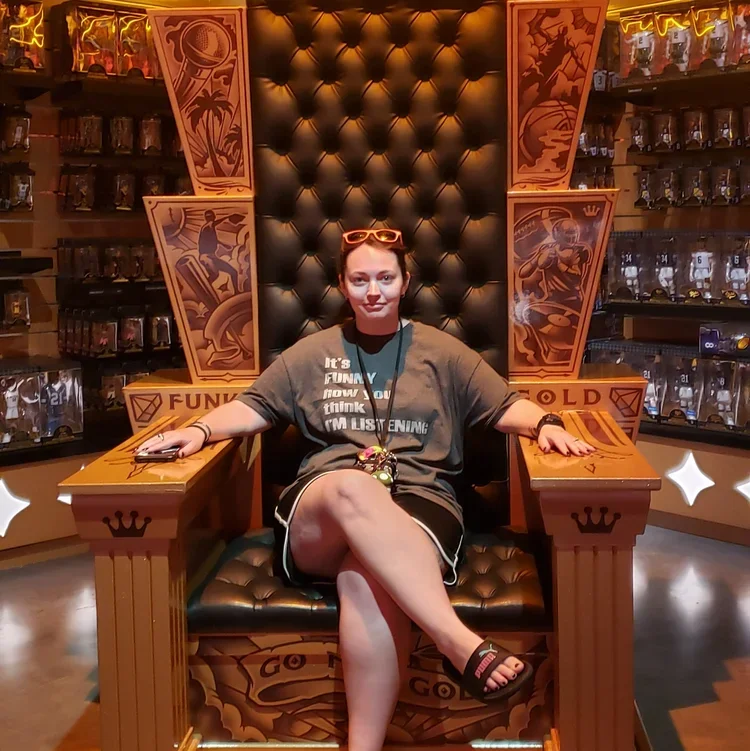 Woman sitting on a large, throne-like chair with a high, tufted backrest, surrounded by shelves of collectibles, with decorative panels and stars on the sides.