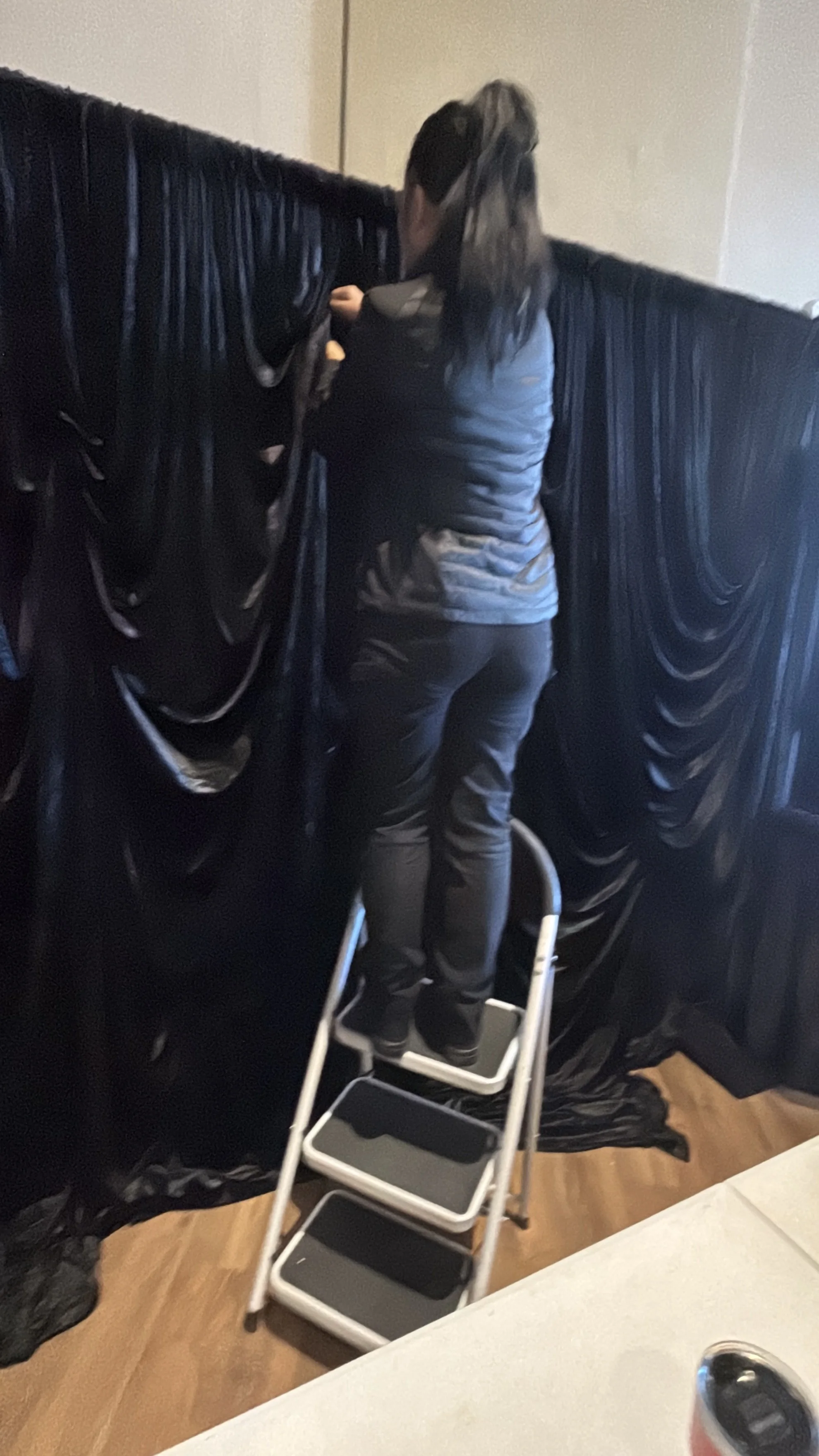 Person standing on a step stool reaching behind black curtains, with a black bag and other items on the floor.