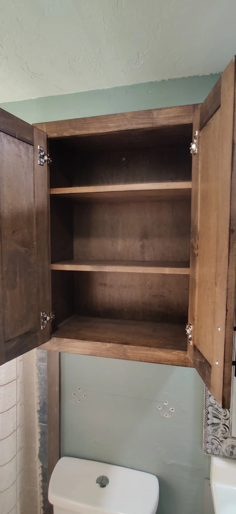 Empty wooden wall cabinet with open doors above a bathroom toilet, with part of a floral curtain visible on the right side.