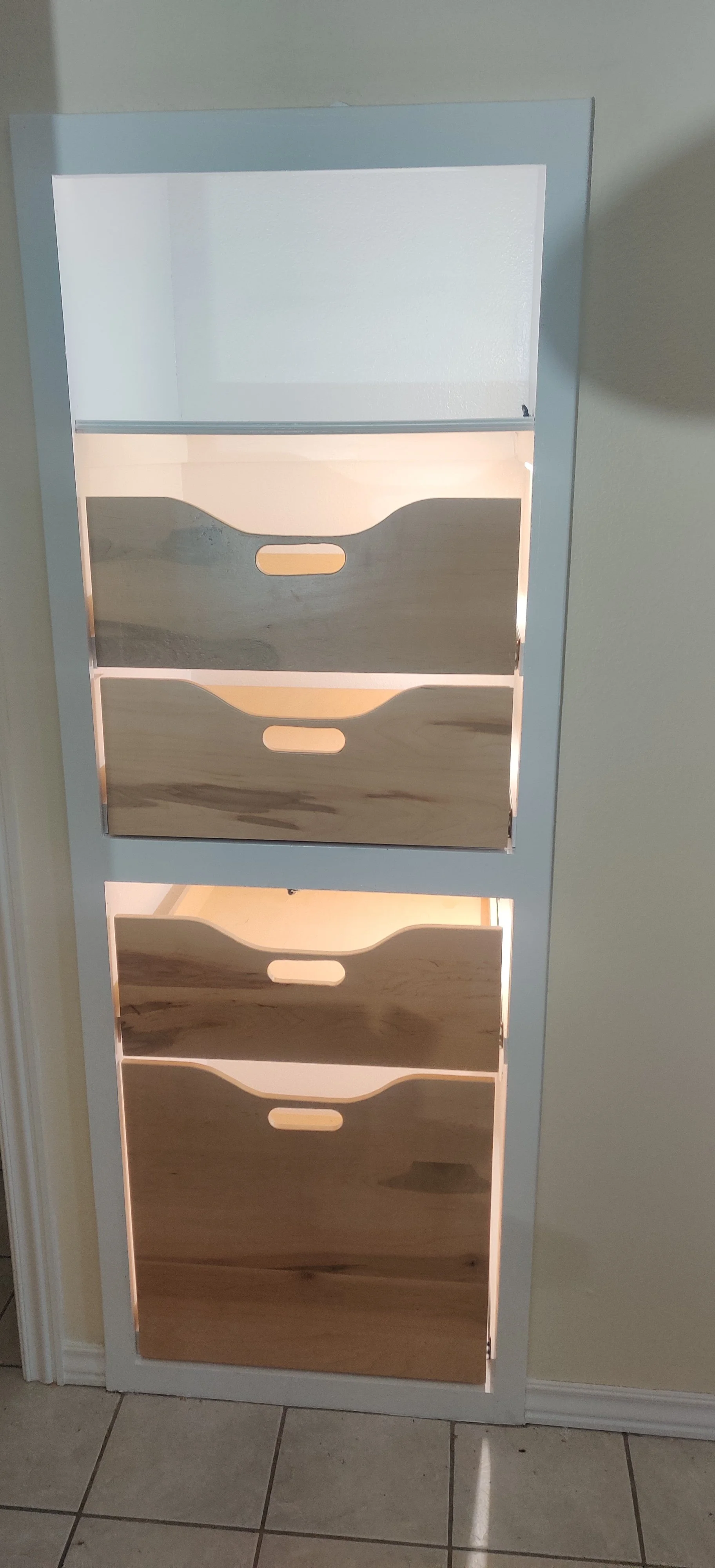 A white cabinet with four shelves featuring sliding wooden doors with cut-out handles, standing against a wall on a tiled floor.