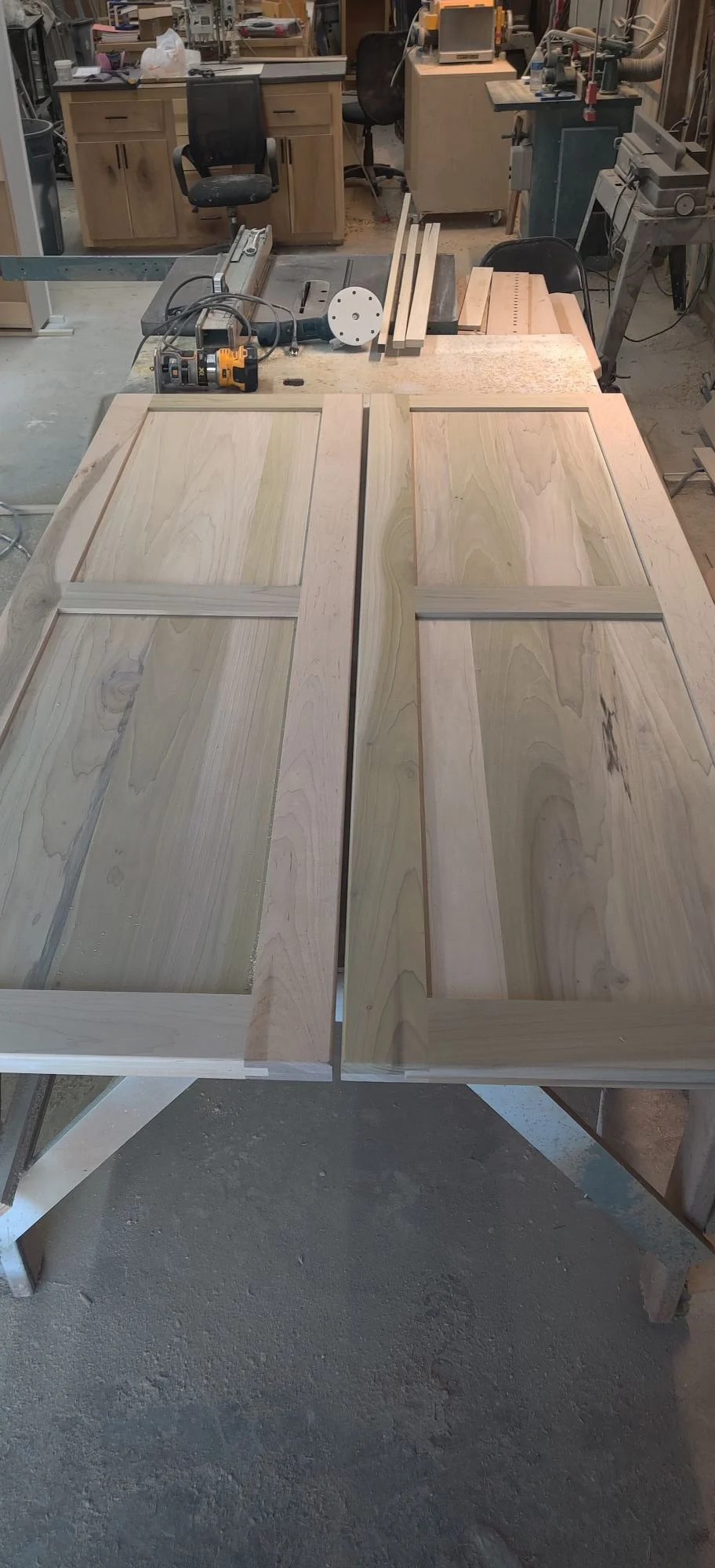 Two wooden cabinet doors lying flat on a workbench in a woodworking shop, with various tools and equipment in the background.