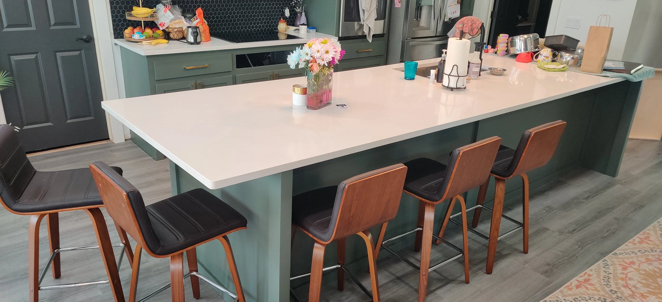 Kitchen with a white island counter, wooden bar stools with black seats, green cabinets, gray wood flooring, and various items on the counter and shelves.