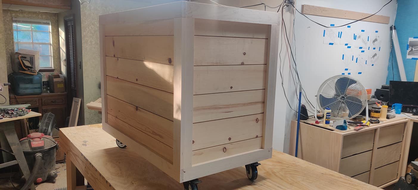 A wooden box on wheels inside a workshop, with tools and equipment visible in the background.