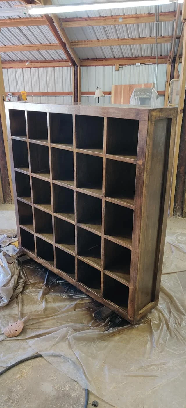 A large wooden shelving unit with multiple cubbyholes inside a workshop or garage, with a metal roof and unfinished walls, placed on plastic sheeting.
