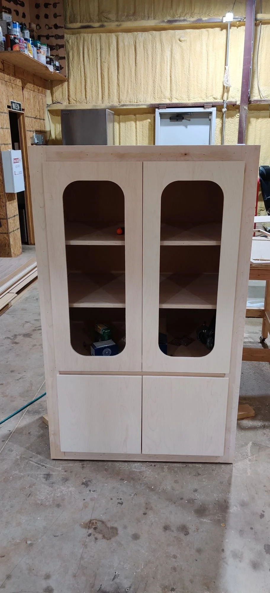 unfinished wooden cabinet with glass-paneled doors inside a workshop.