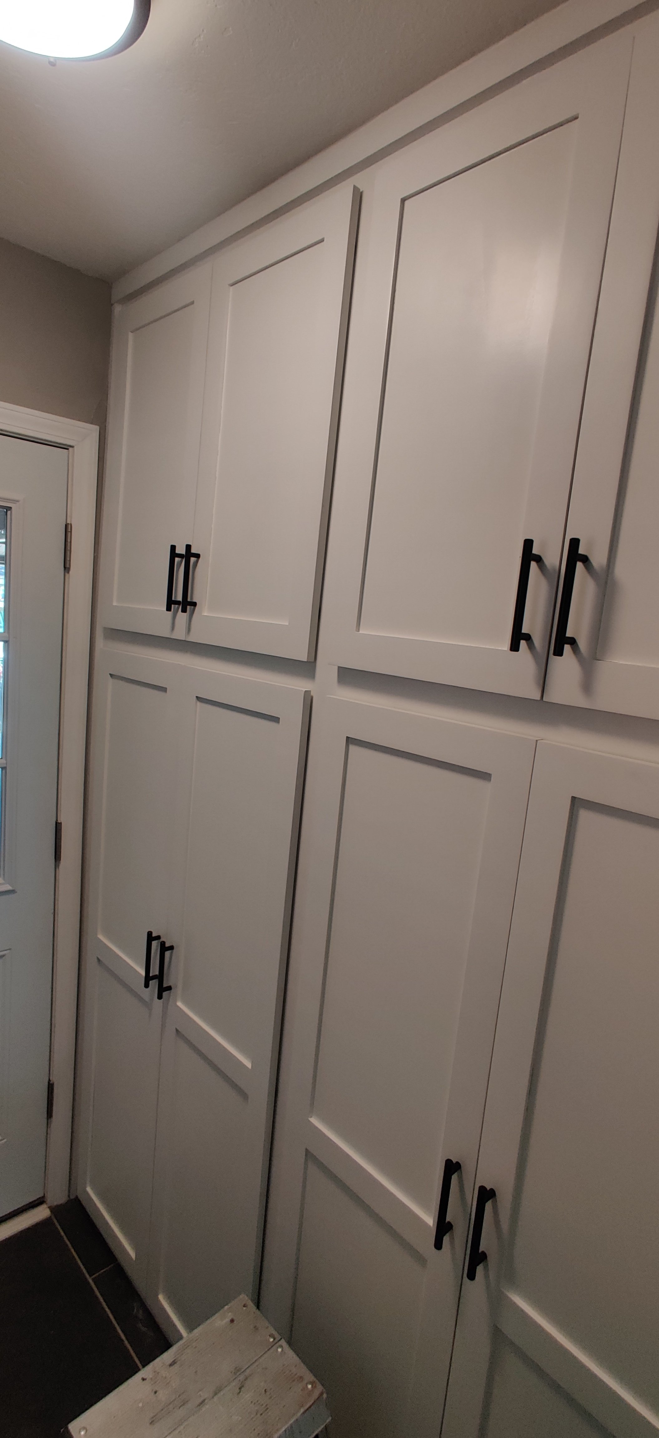 Photo of white kitchen cabinets with black handles, next to a door with a window, and a small wooden step stool on a black tile floor.