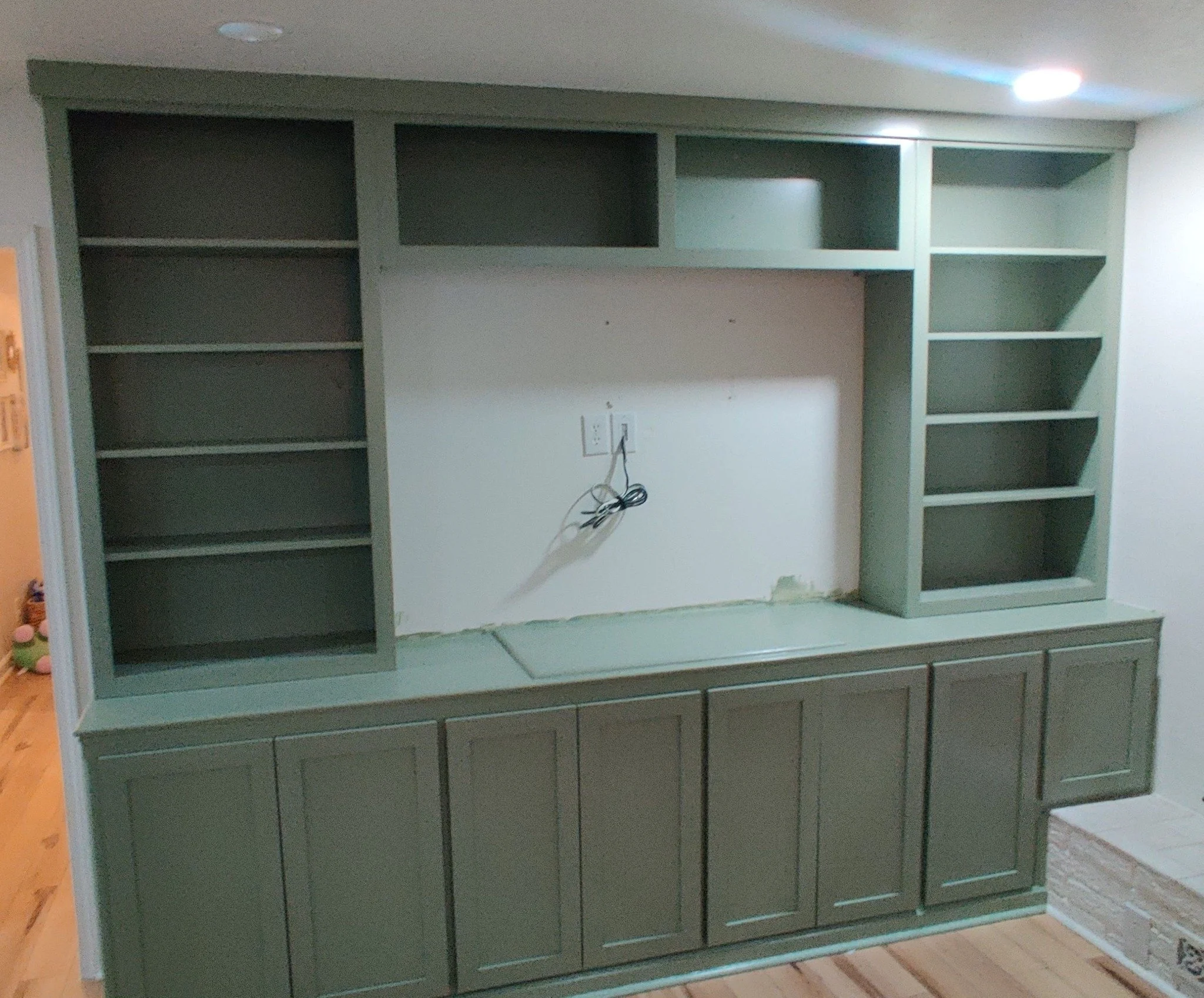 Gray built-in bookshelf with multiple empty shelves, situated in a room with a white wall, electrical outlets, and an electrical cord hanging from the middle shelf.