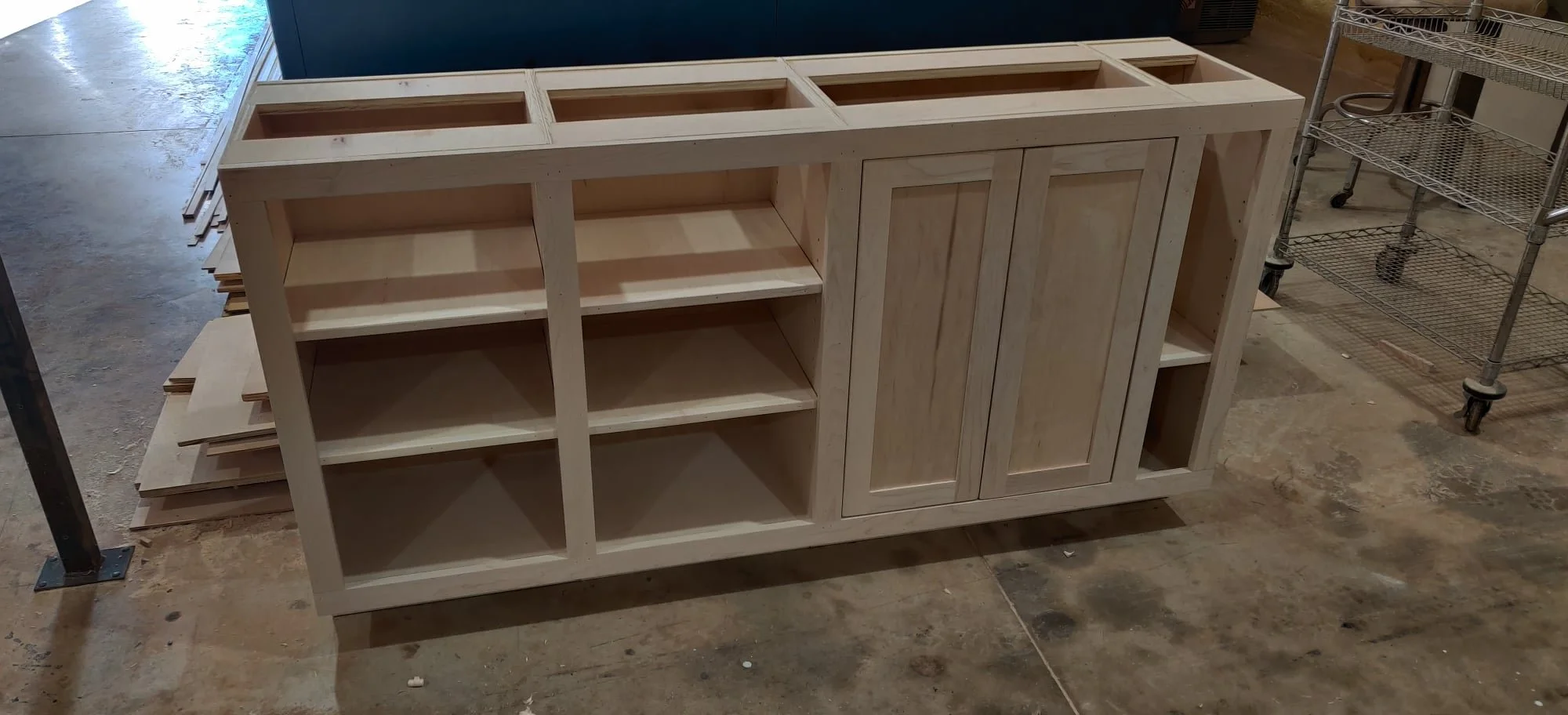 Unfinished wooden cabinet with open shelves and two doors, situated on a concrete floor in a workshop.