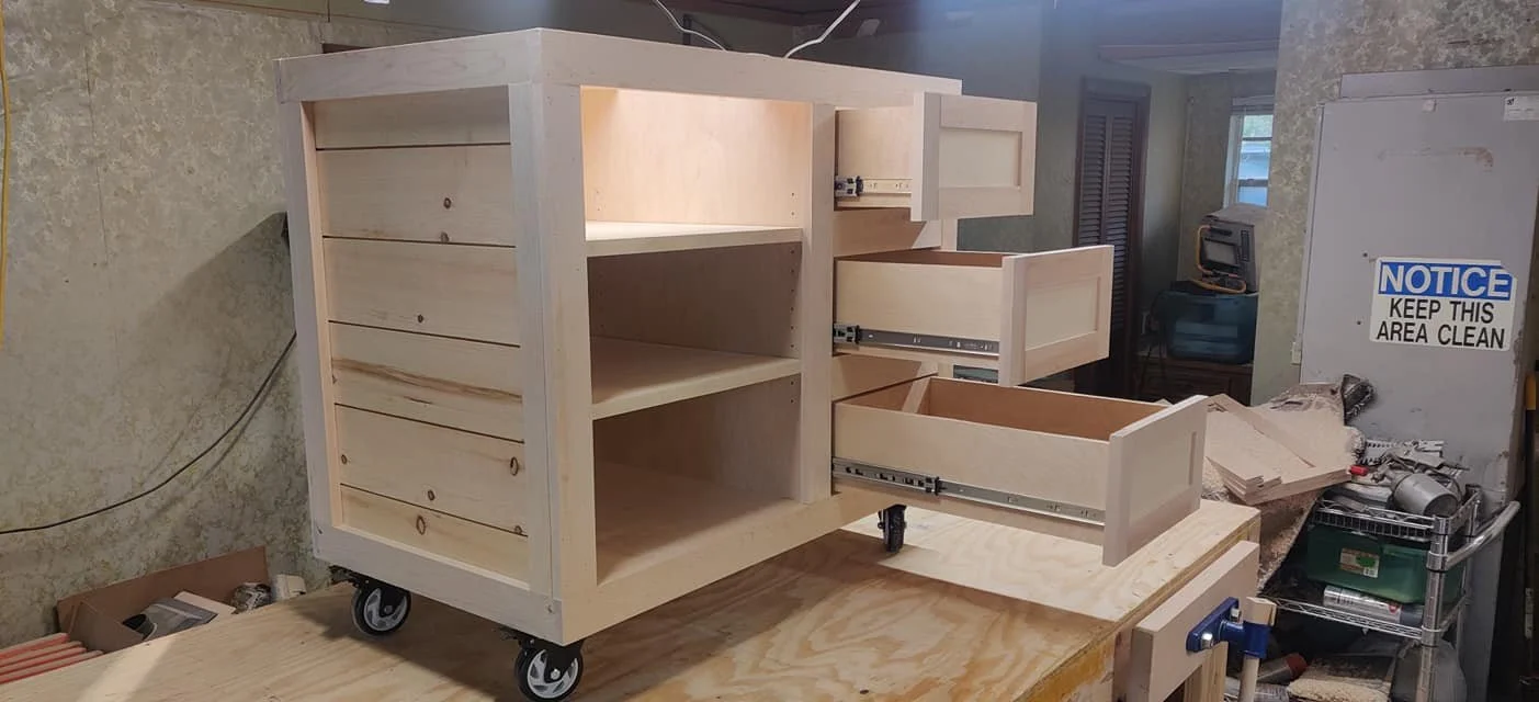 Wooden furniture piece on caster wheels in a woodworking shop, with drawers open and construction tools nearby.