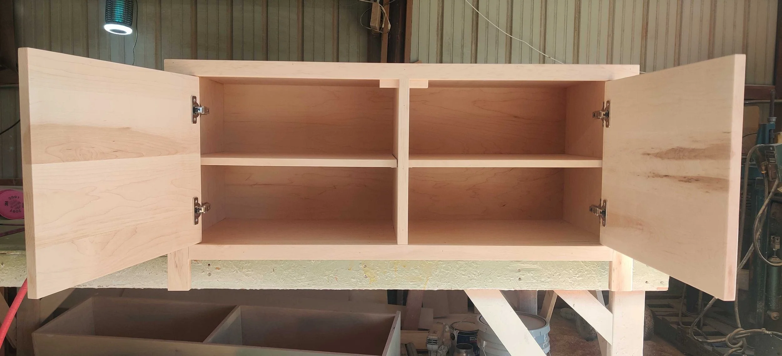 Unfinished wooden cabinet with four compartments and open doors, placed on a workbench in a workshop.