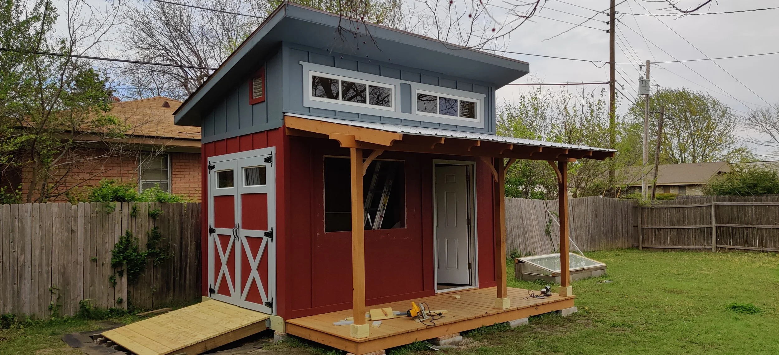 A small two-story playhouse or shed under construction in a backyard with a wooden deck, a ramp, and a partially finished exterior in red and blue with white trim.