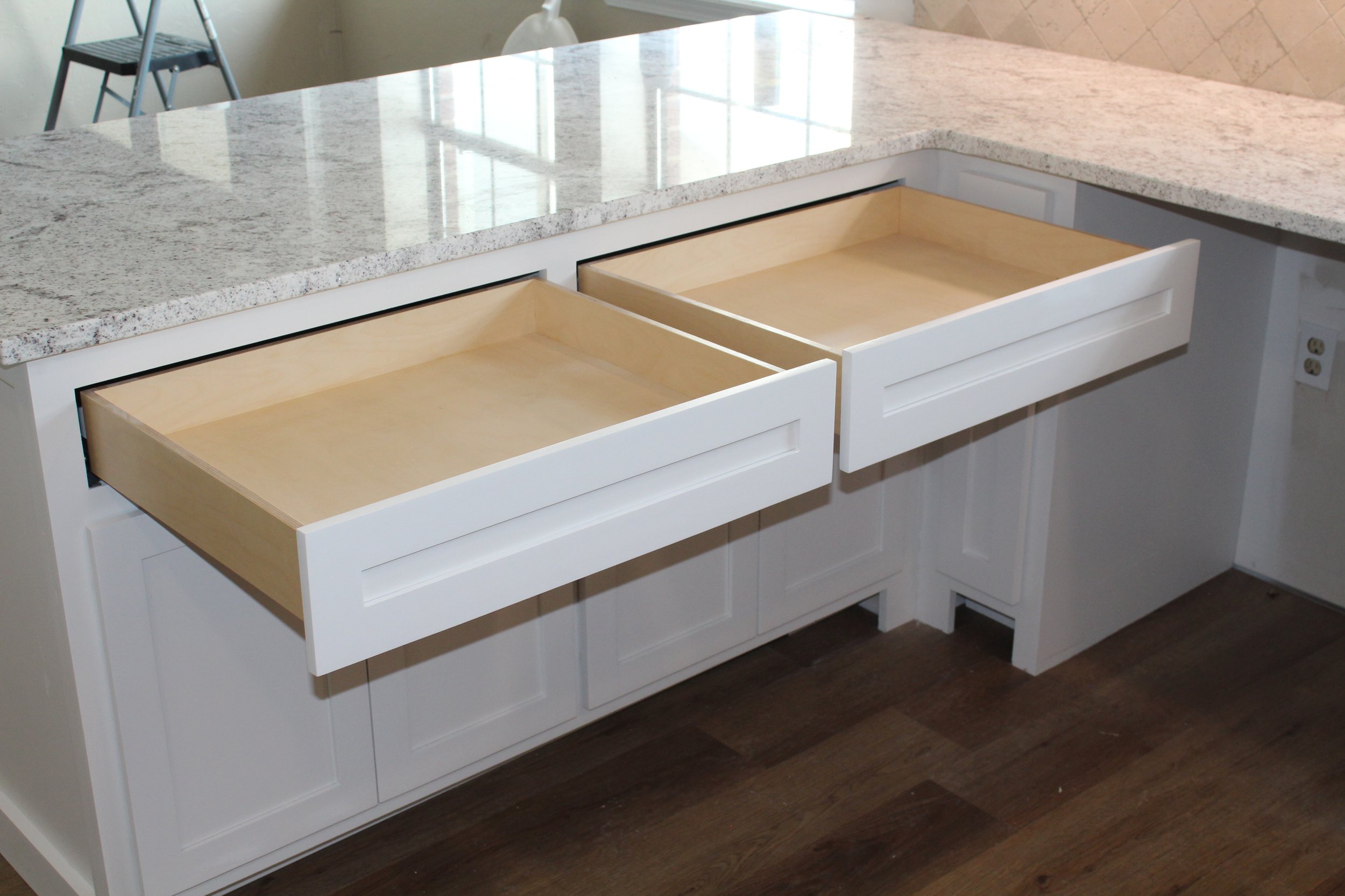 A kitchen island with a white base and two open drawers, a granite countertop, and wooden interior drawers. In the background, there is a black and silver step stool and a white lamp.