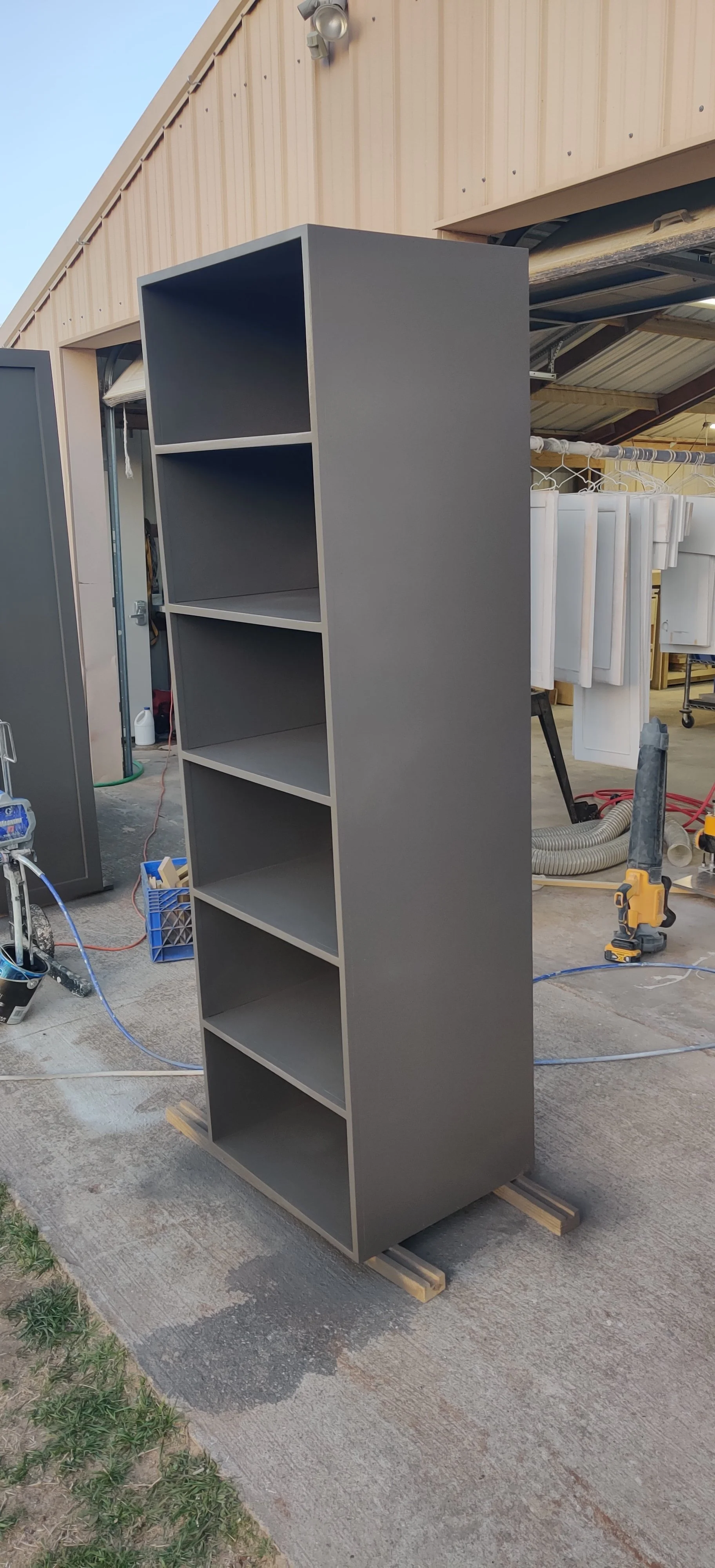 A tall, gray bookshelf with six open shelves standing outdoors on a concrete surface, with a building in the background and construction tools around.