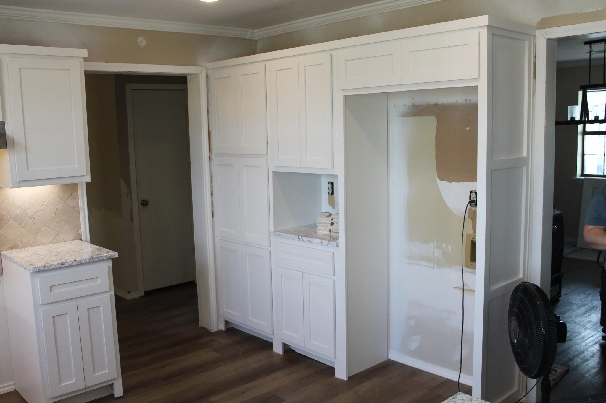 Living room with white built-in cabinets and a partial kitchen counter, under renovation. Walls are beige, with some patches, and hardwood floors.