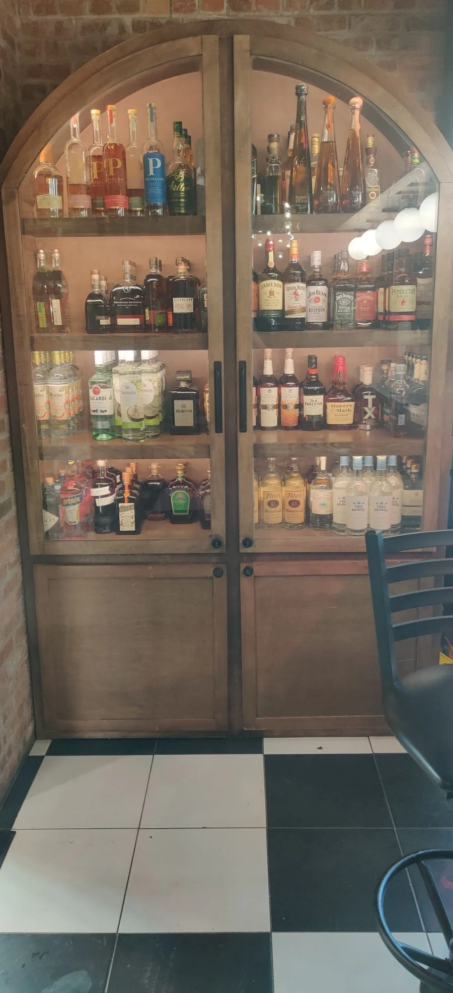 A wooden bar cabinet filled with various bottles of alcohol, set against a brick wall, with a black and white tiled floor and part of a chair visible on the right.