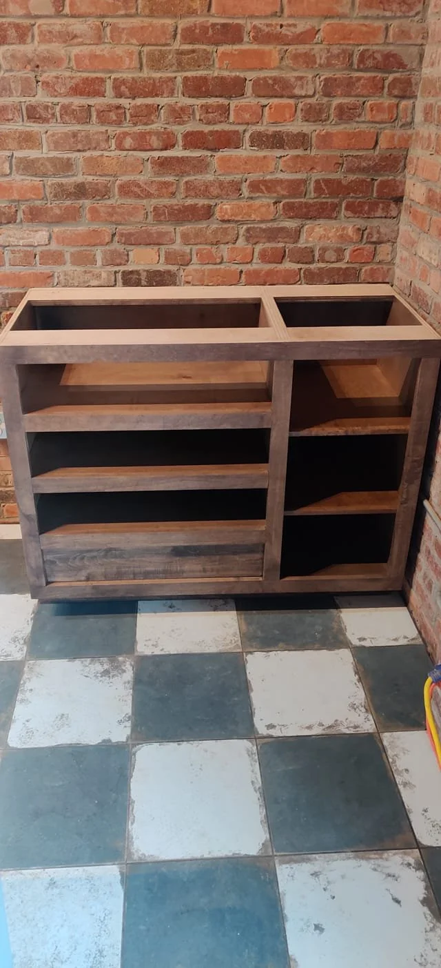 Unfinished wooden cabinet with open shelves placed against a brick wall, on a floor with black and white tiles.