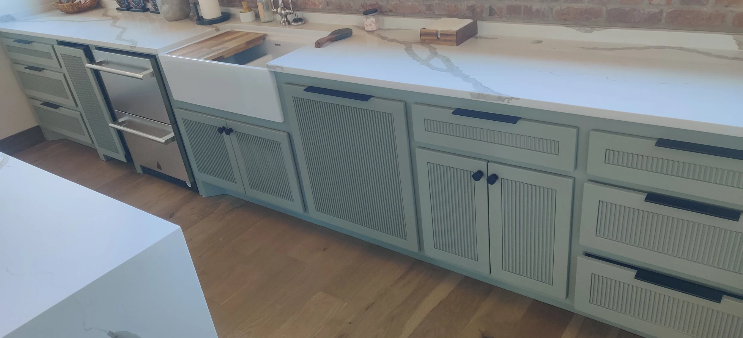 Kitchen countertop with a farmhouse sink, wood cutting board, and various bottles and containers. The cabinets below have a light blue-green color with black handles, and the floor is wood. Parts of the wall show exposed brick and unfinished areas.