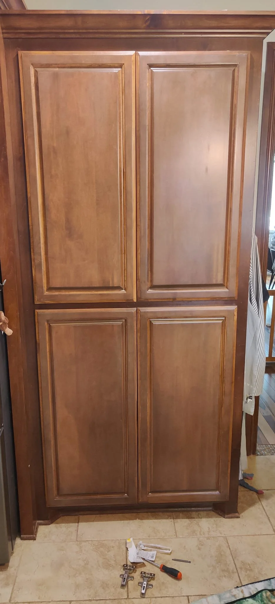 Unfinished wooden cabinet with four doors, missing hardware, in a home kitchen. Tools and hardware pieces on the tile floor in the foreground.