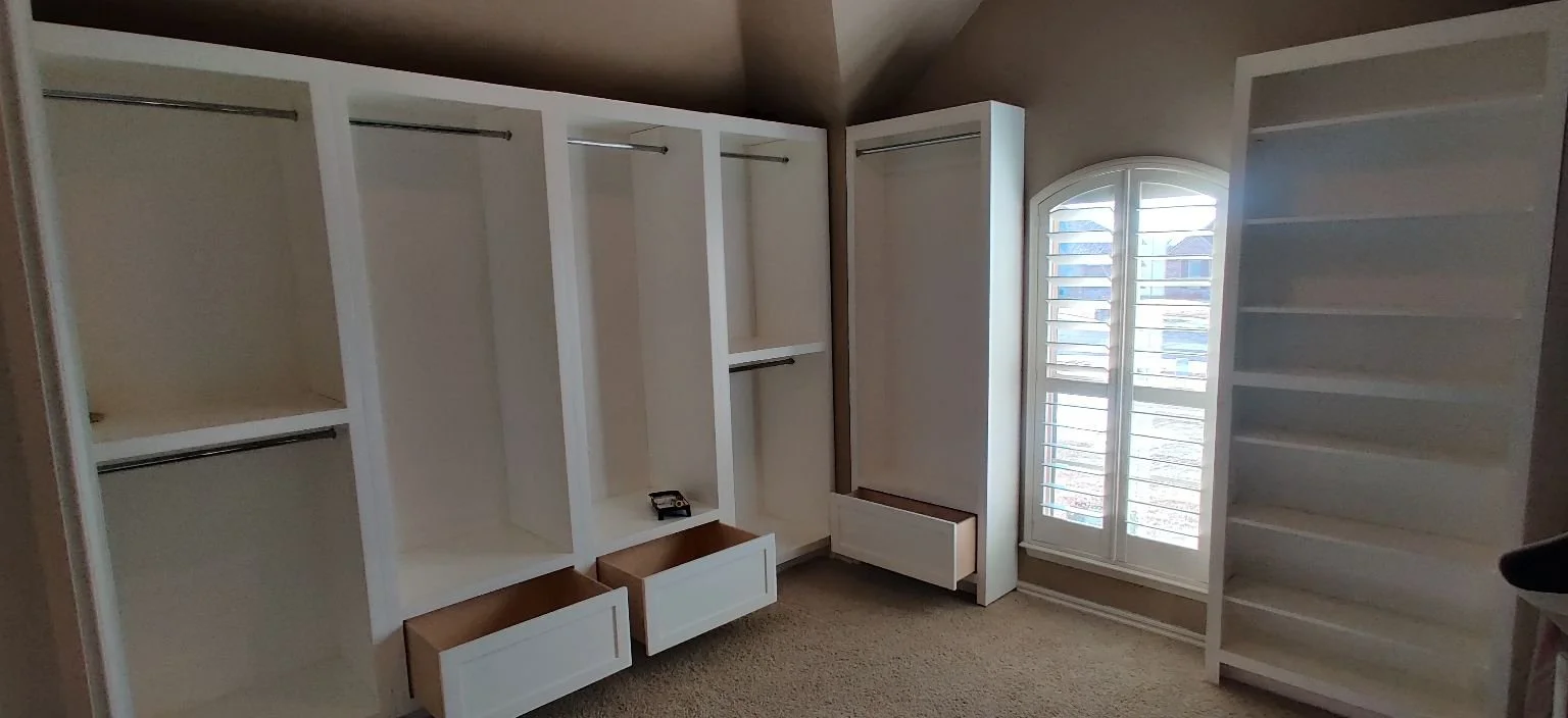 Empty white built-in wardrobe with multiple open compartments and drawers, next to a window with shutters in a beige room.