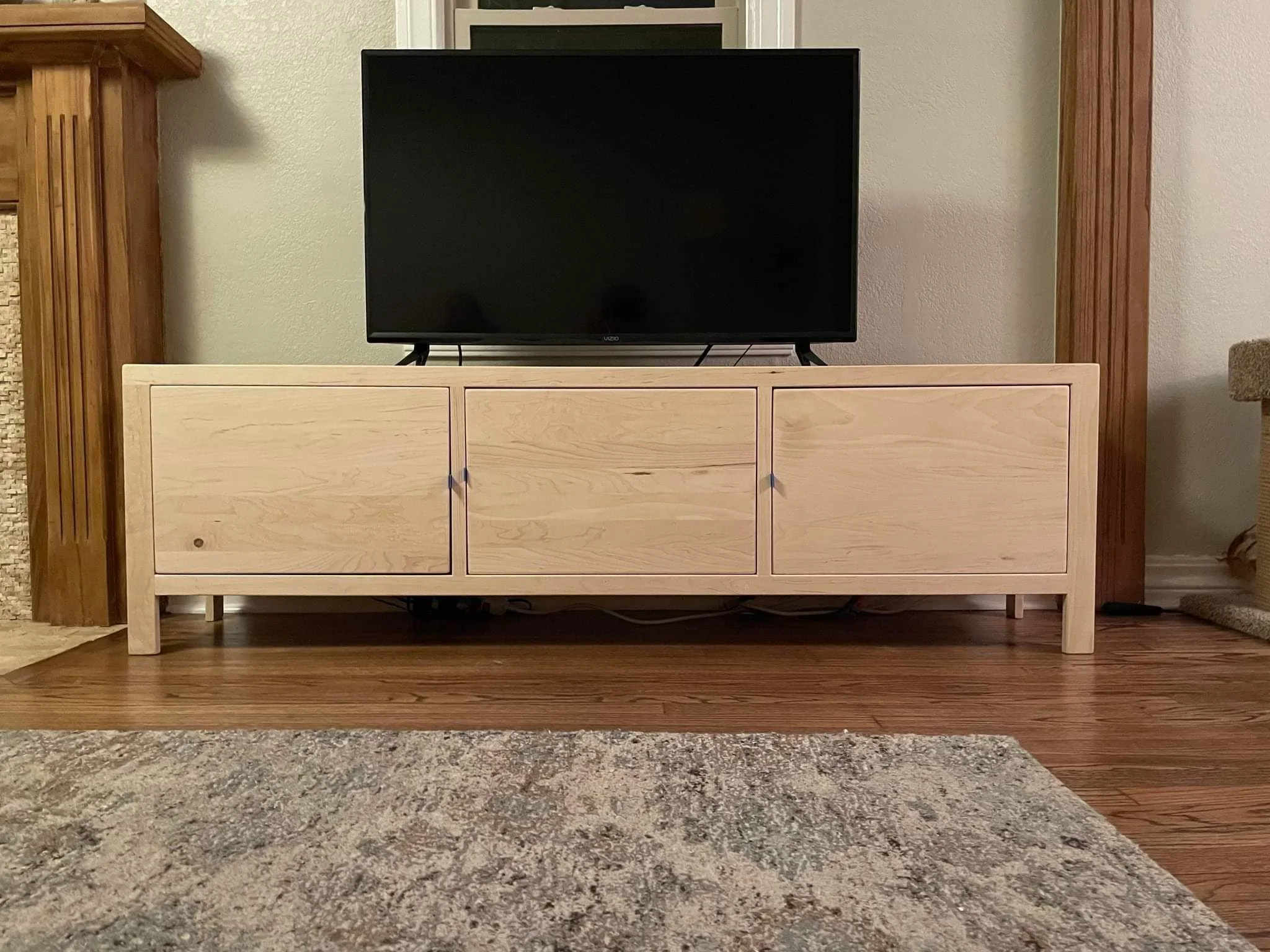 Empty black television screen on a wooden TV stand against a beige wall, with a wooden cabinet on the left and a fireplace in the background.