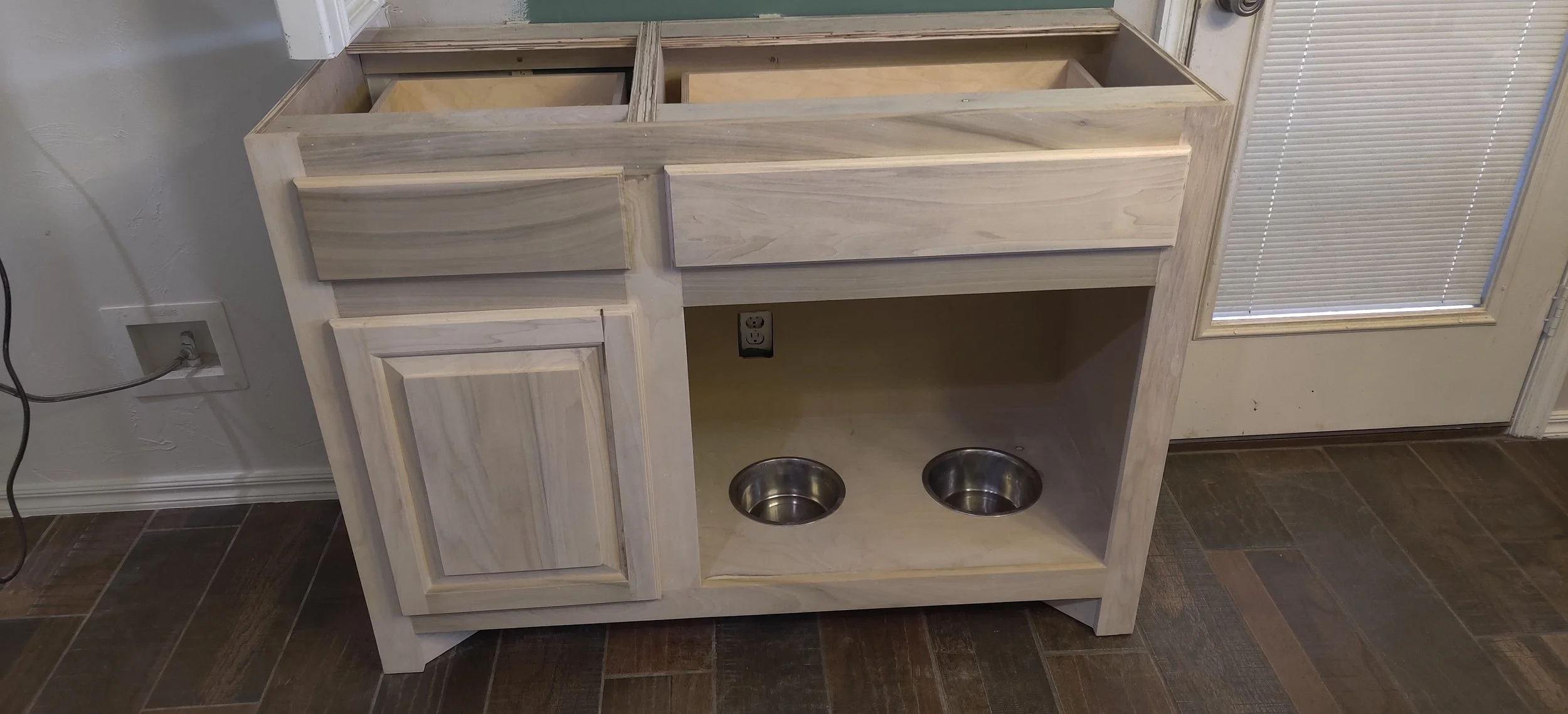 Unfinished wooden dog feeding station with two stainless steel bowls, a cabinet, and an open space for a dishwasher or sink, situated in a room with a window and a tile floor.