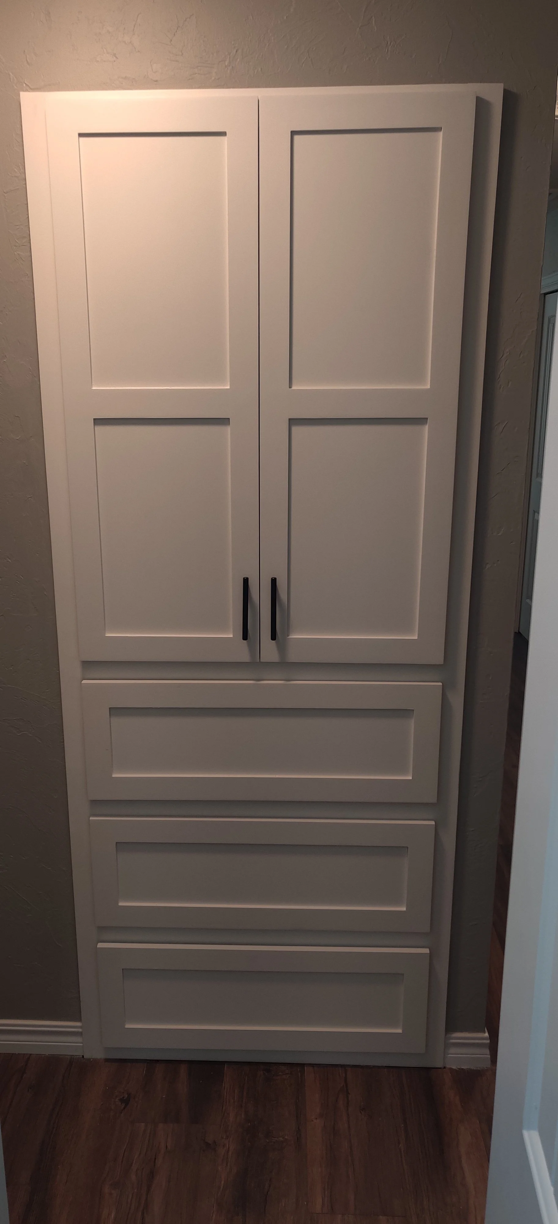 White built-in cabinet with two upper doors, two middle drawers, and two lower drawers, all with black handles, mounted on a beige wall in a room with wood flooring.