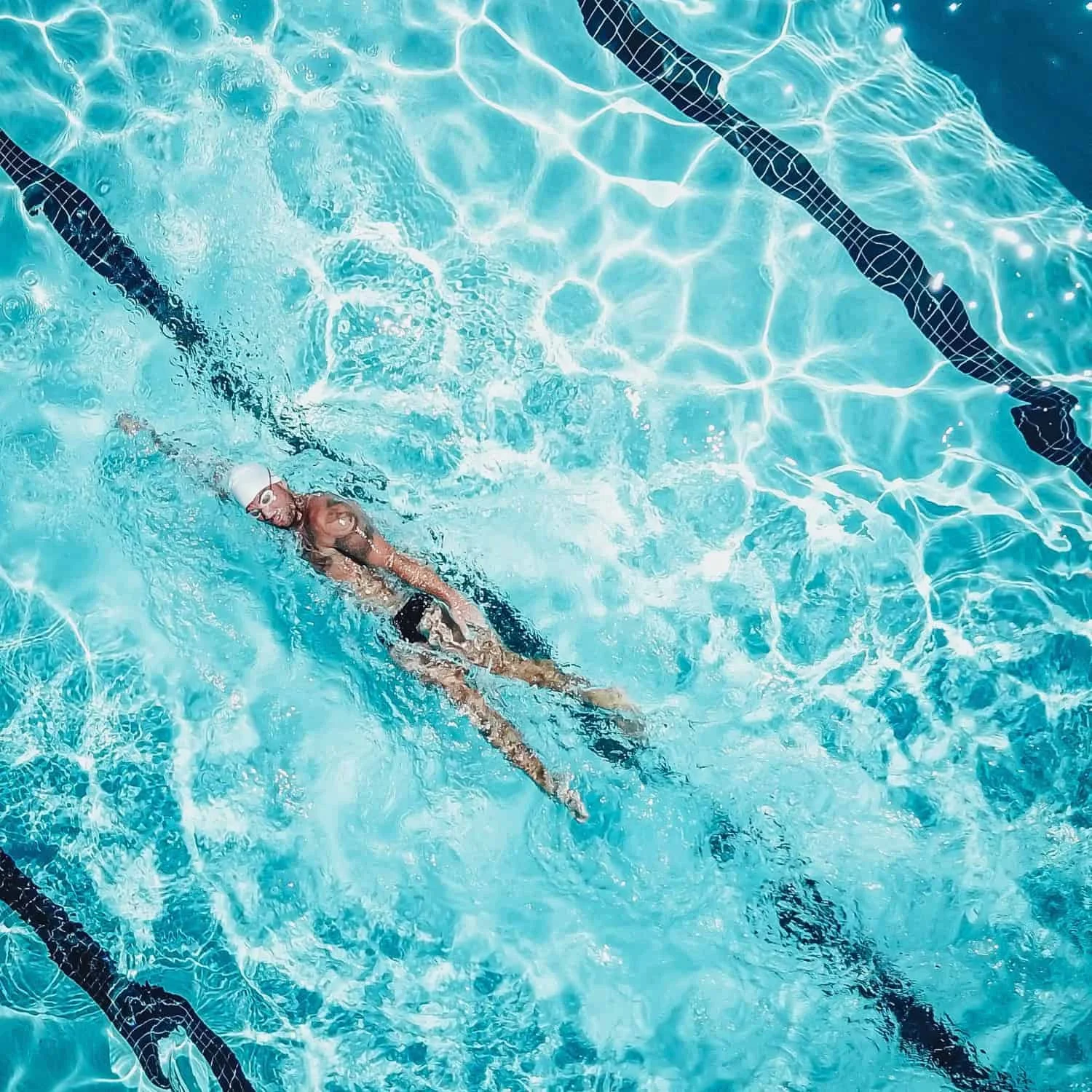 A swimmer in a swimming pool wearing a white swimming cap and goggles, swimming in the water near the pool lane divider.