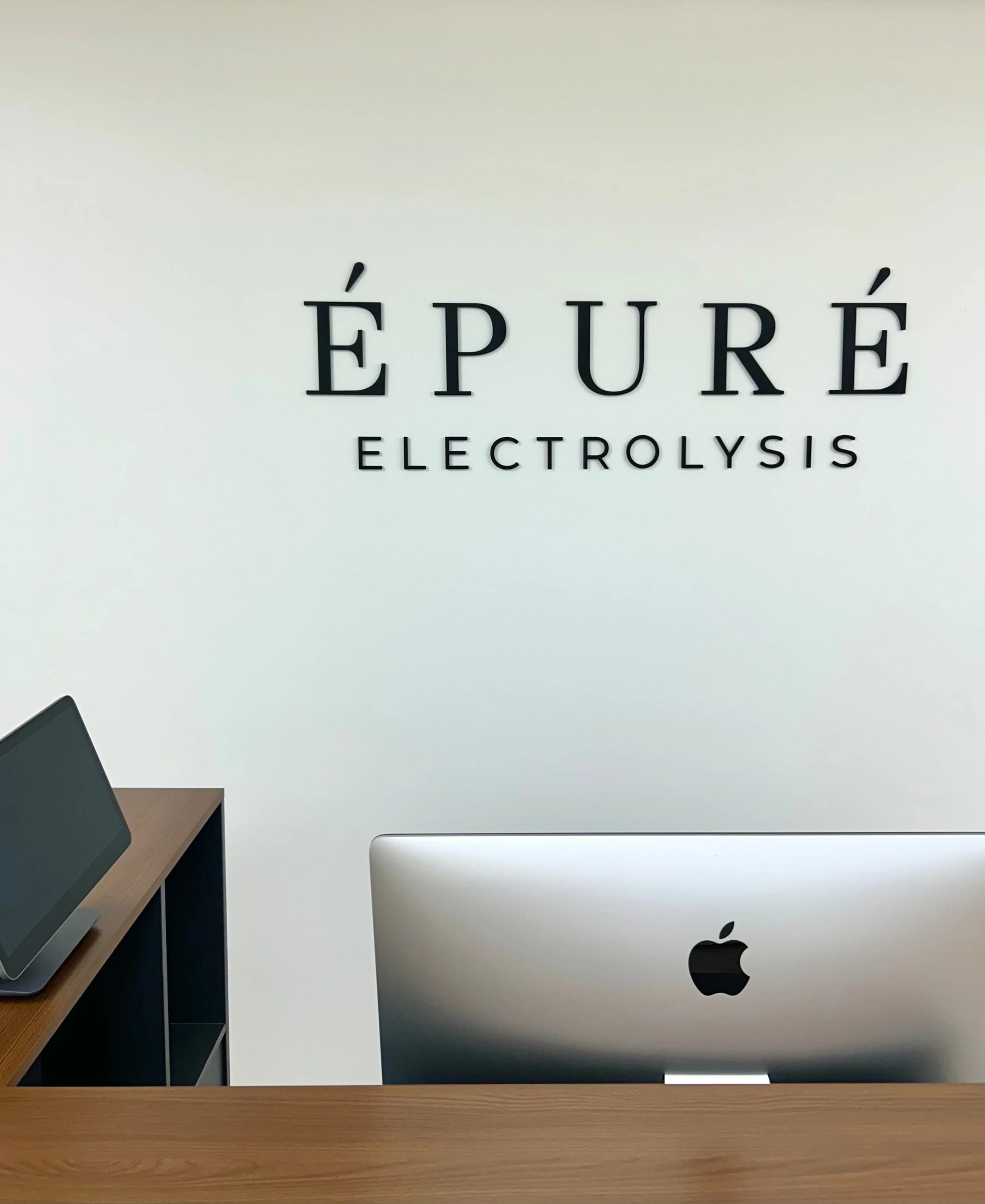 Desk with Apple MacBook and an iPad on a wooden surface, against a white wall with 'ÉPURE ELECTROLYSIS' written on it.