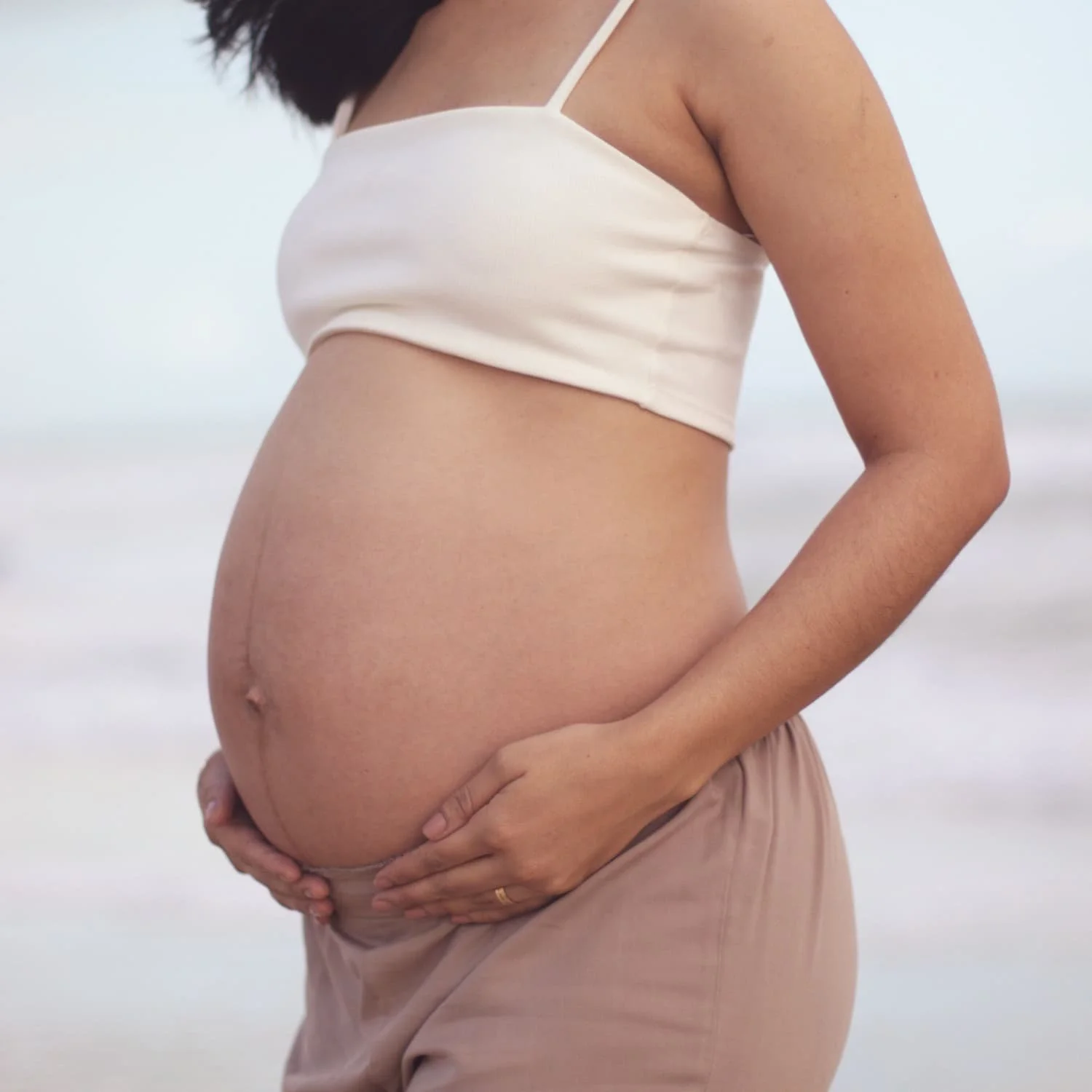 Close-up of a pregnant woman holding her belly on a beach, wearing a white tank top and beige pants.
