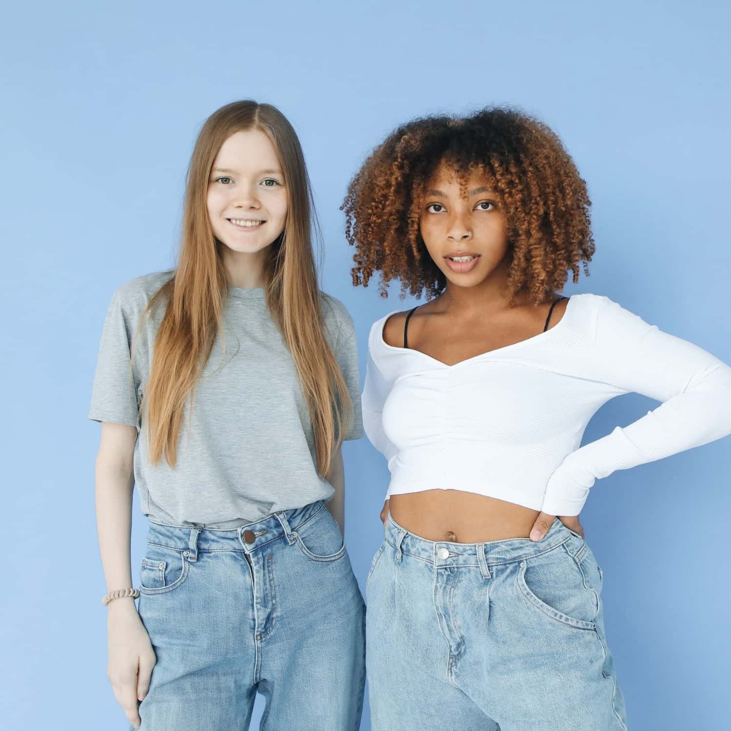 Two young women standing against a blue background, one with straight red hair wearing a gray t-shirt and jeans, and the other with curly hair wearing a white crop top and jeans, posing confidently.