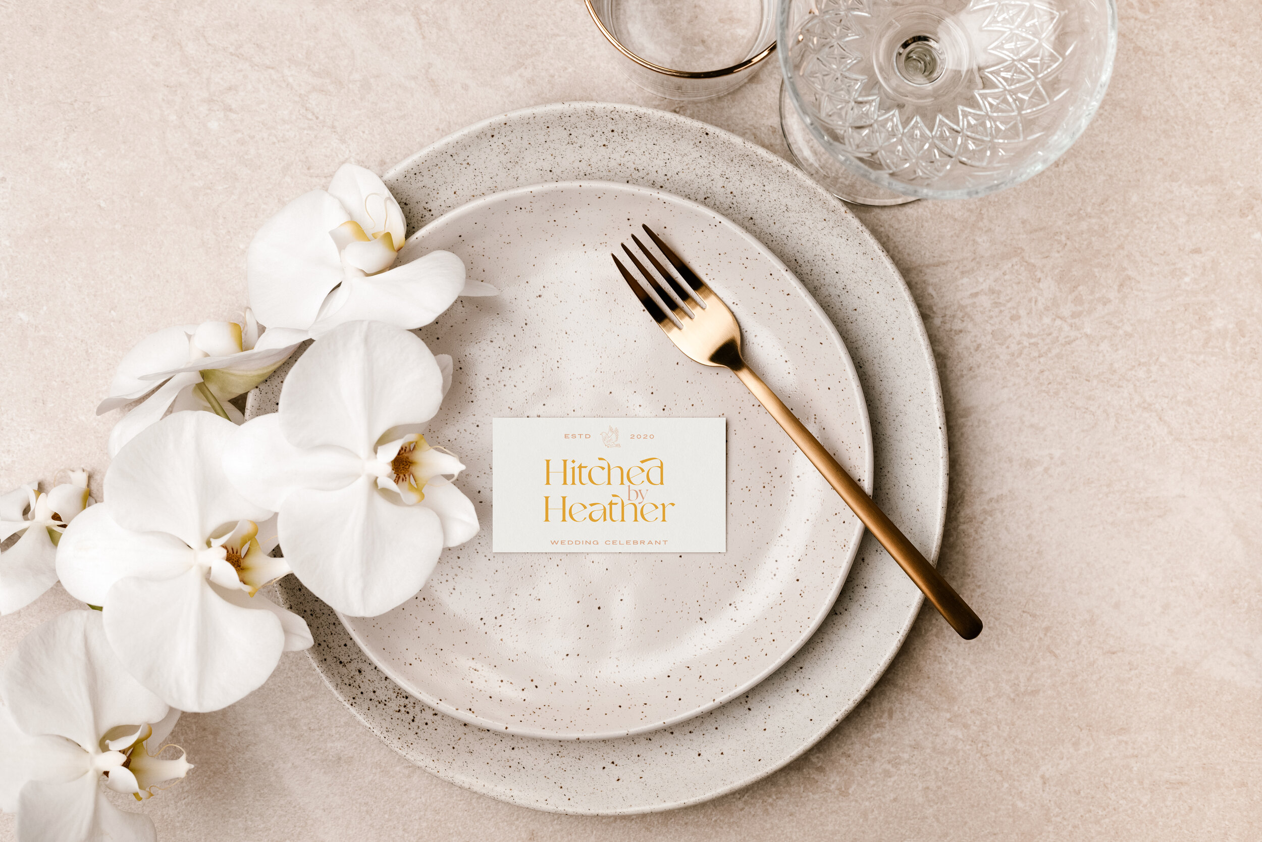 A wedding place setting with a white speckled plate, a gold fork, white orchids, and a card reading 'Hitched by Heather' on a beige textured surface.