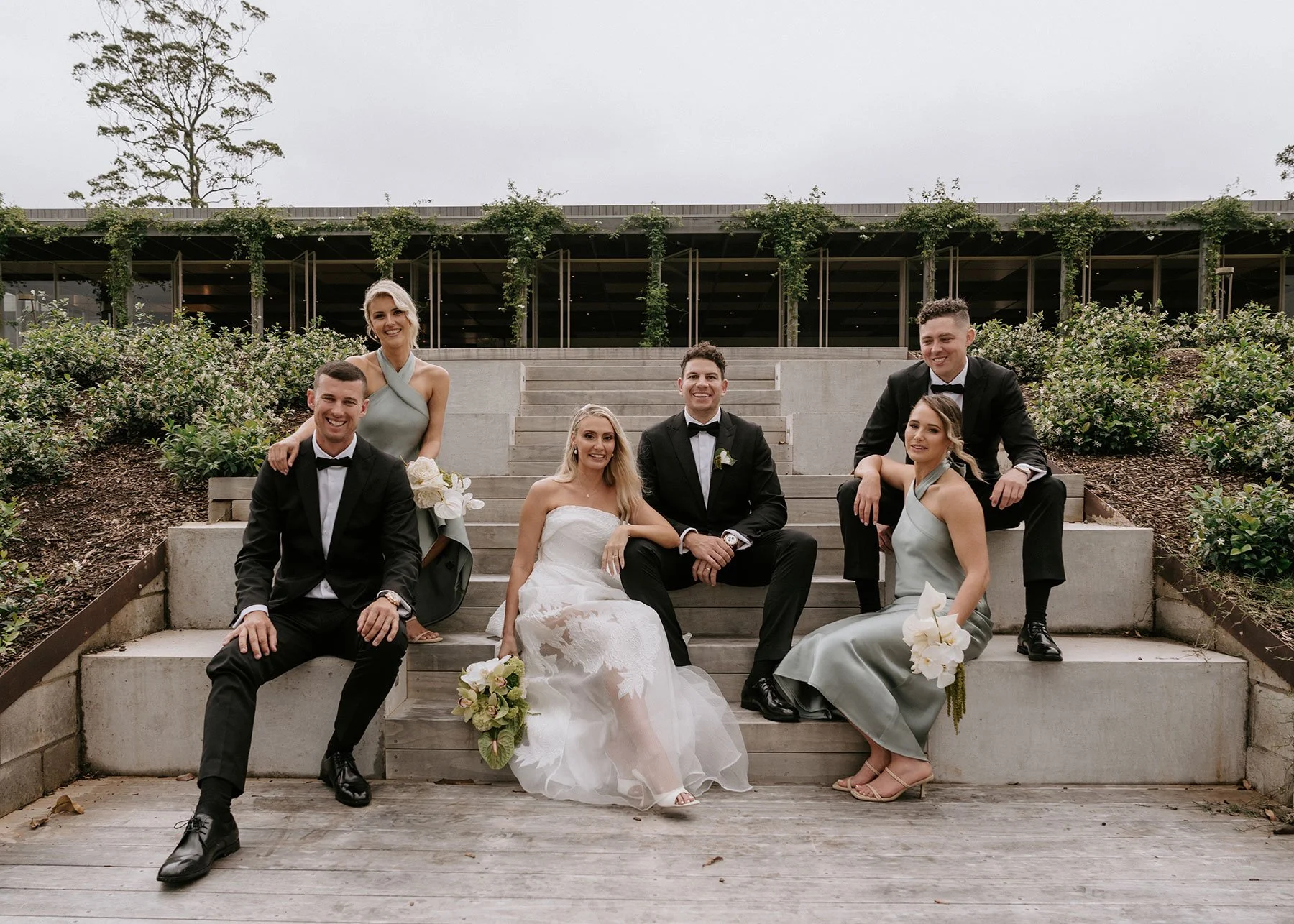A wedding party sits on the steps outdoors, including a bride in a white gown, groomsmen in tuxedos, and bridesmaids in matching dresses, surrounded by greenery.