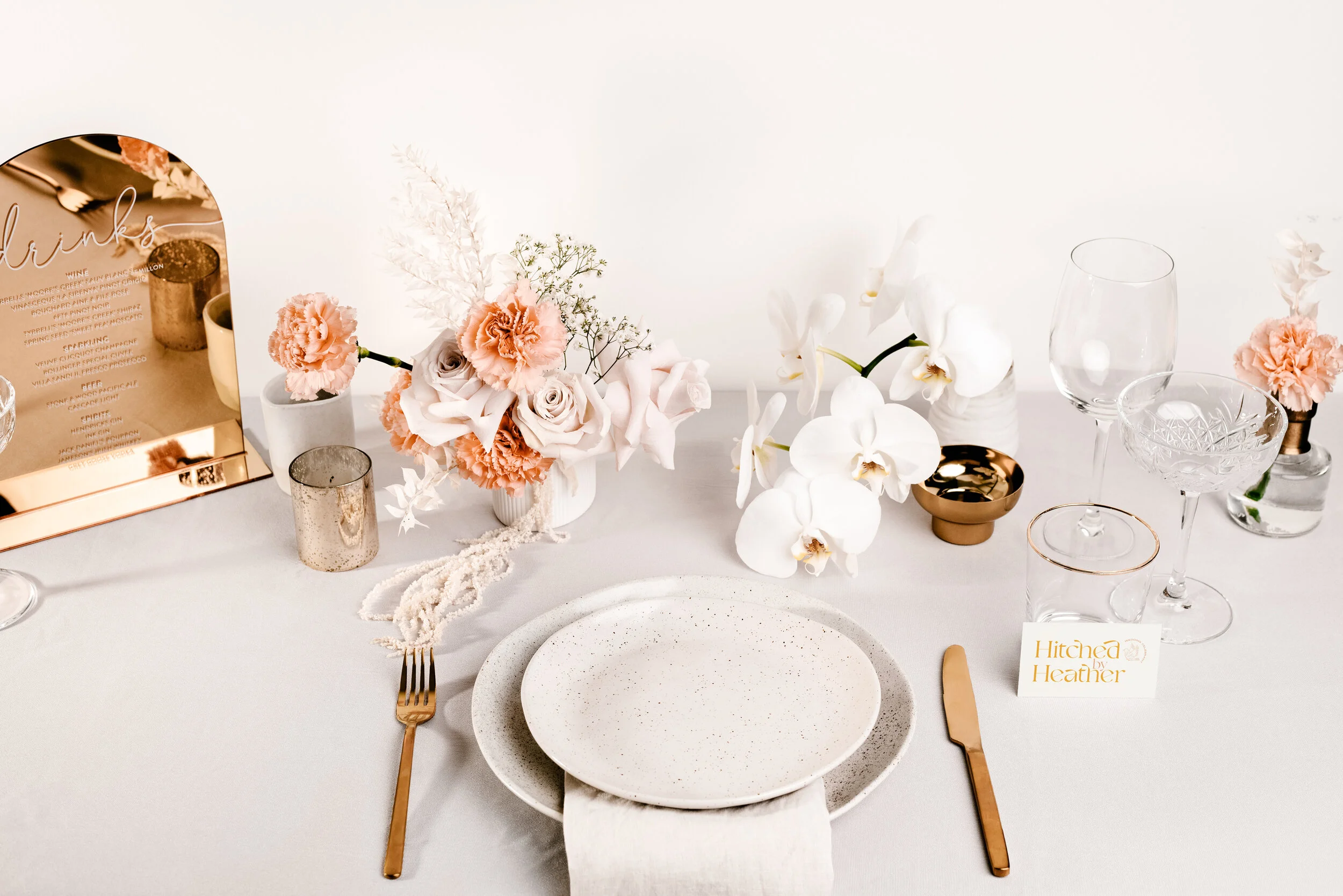 Elegant table setting with white and peach flowers, glassware, and gold cutlery, with a menu card on the side.