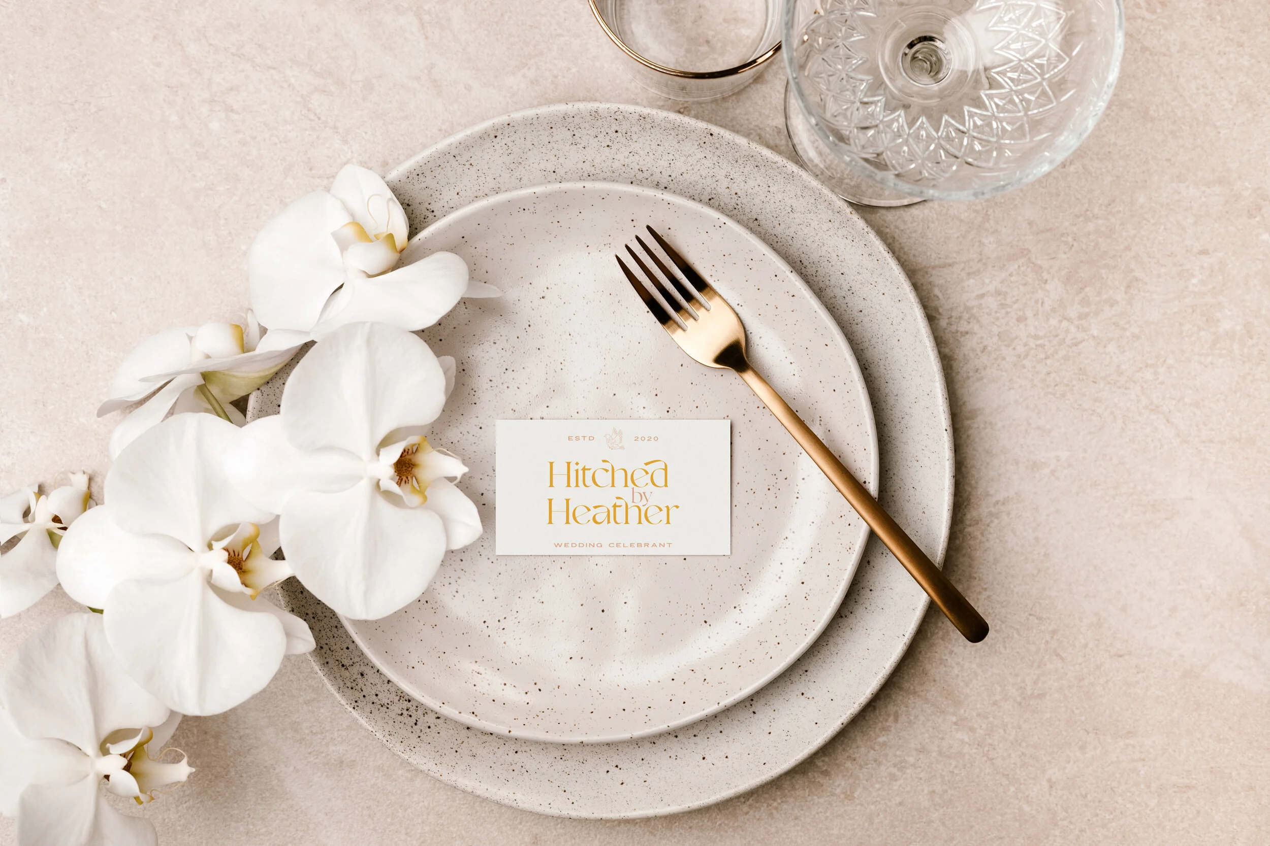 Elegant table setting with stacked white speckled plates, a gold fork, white orchids, a wine glass, and a card reading "Hitched by Heather" on a light beige background.