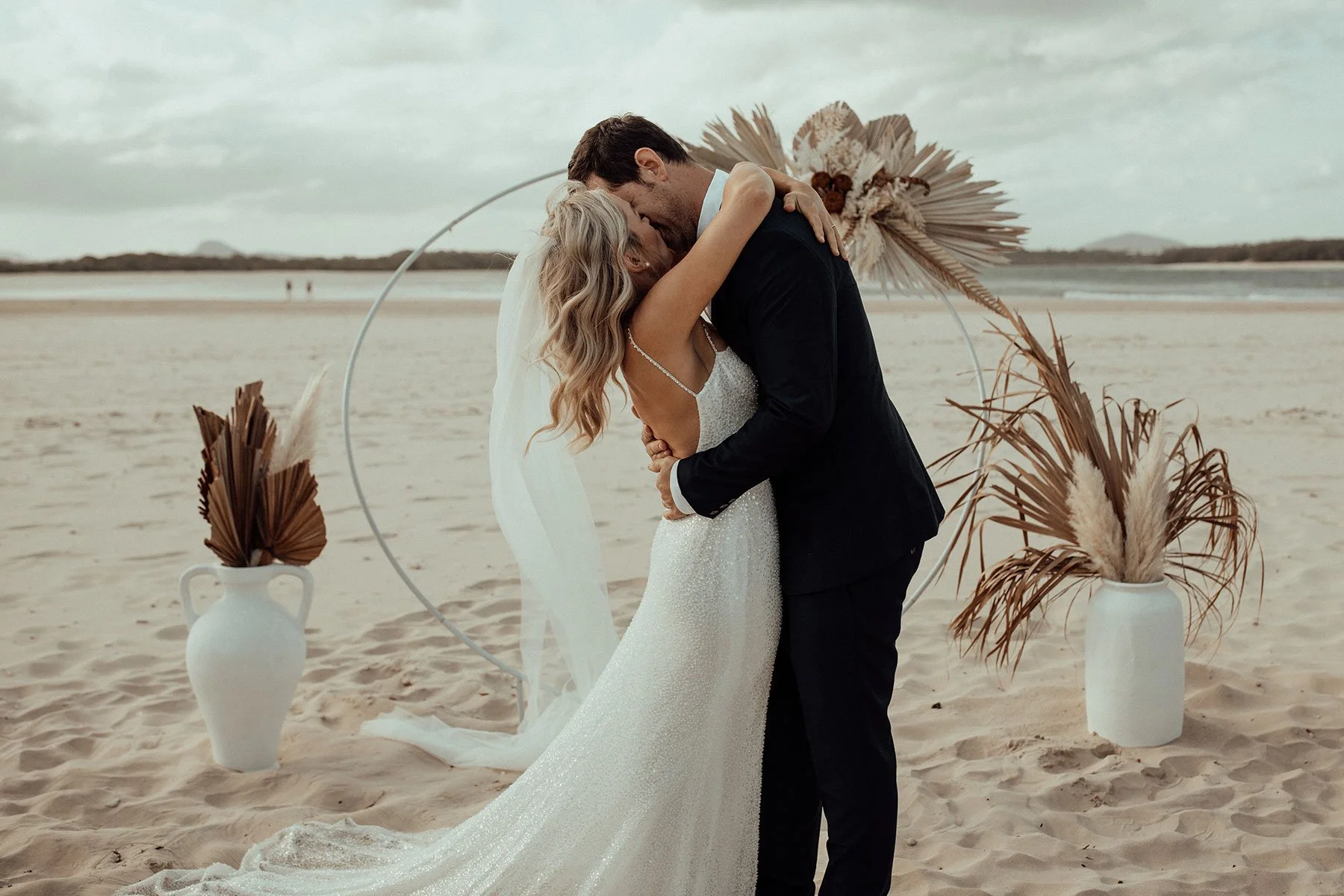 A bride and groom kissing on a sandy beach during their wedding ceremony, surrounded by decorative vases with dried palm fronds and a floral arch.