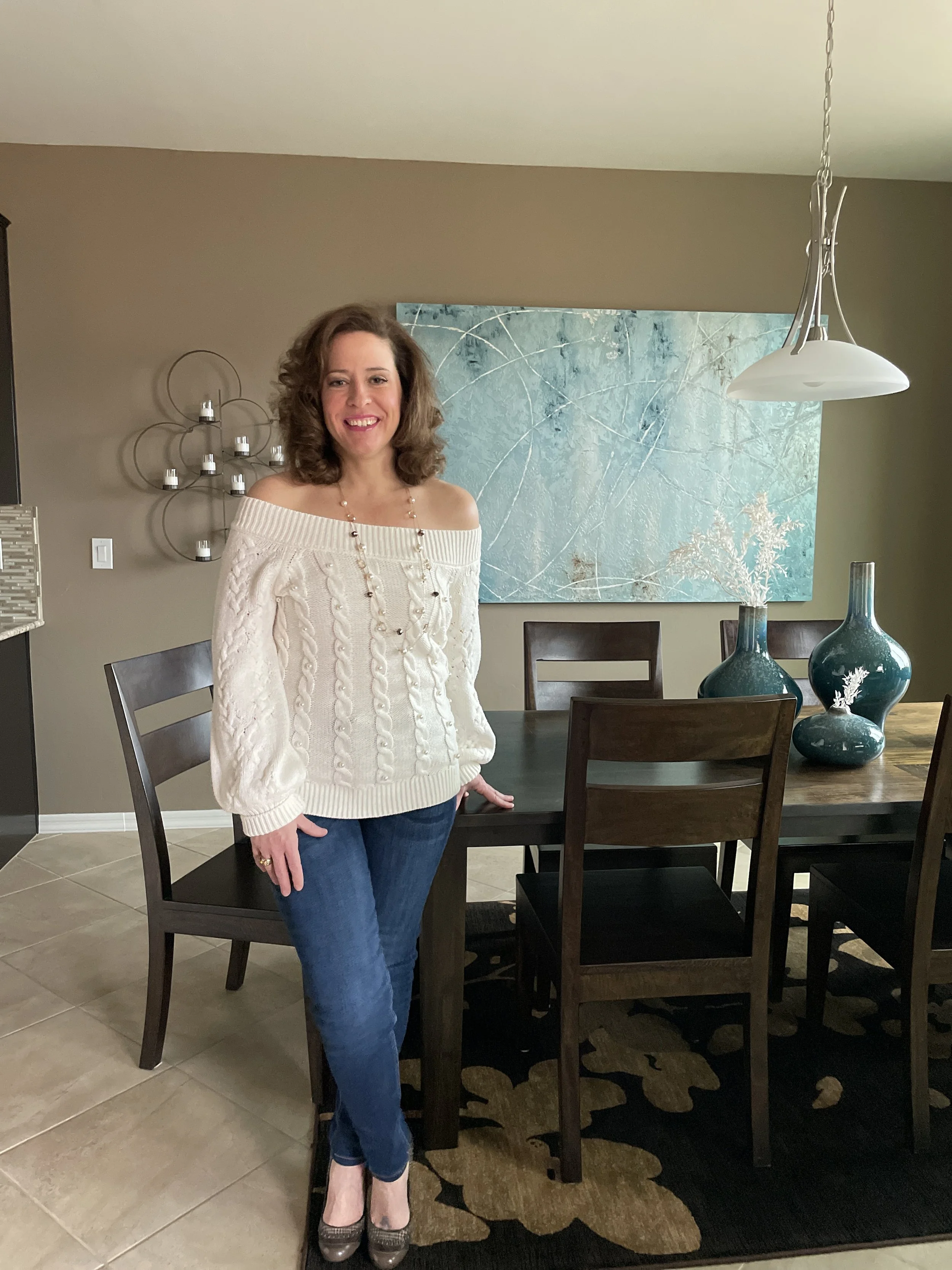A woman with curly brown hair wearing a white off-shoulder sweater, blue jeans, and high heels, standing in a dining room with her hand on a table. Behind her is a wall with abstract art and a decorative metal wall sculpture. The room has a wooden dining table, dark chairs, and decorative vases on the table.