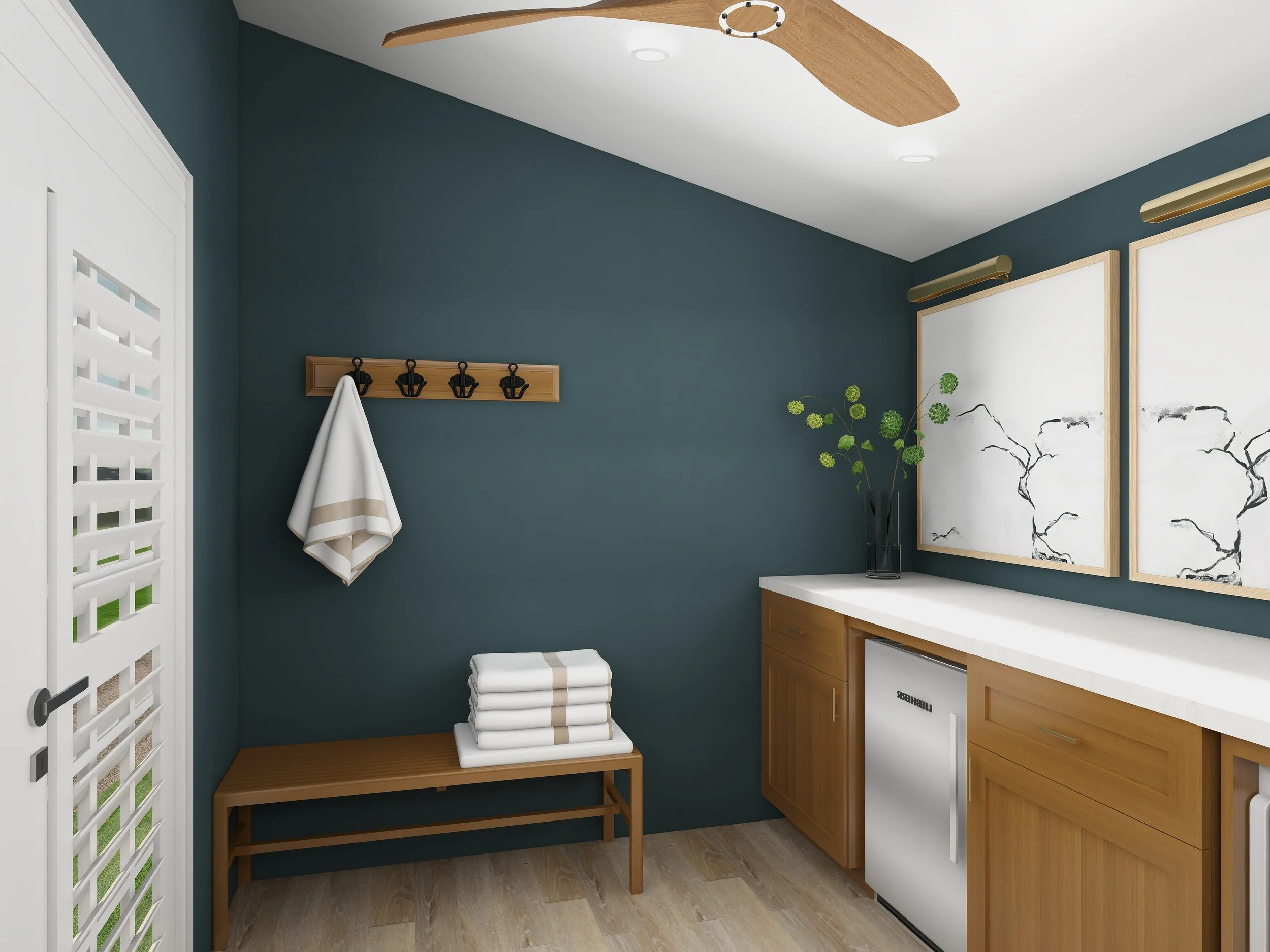 A modern laundry room with green walls, a wooden bench with neatly folded towels, a wall-mounted coat rack with hooks, a small countertop with a cabinet, a mini fridge, and decorative wall art featuring black ink sketches of trees. A ceiling fan with
