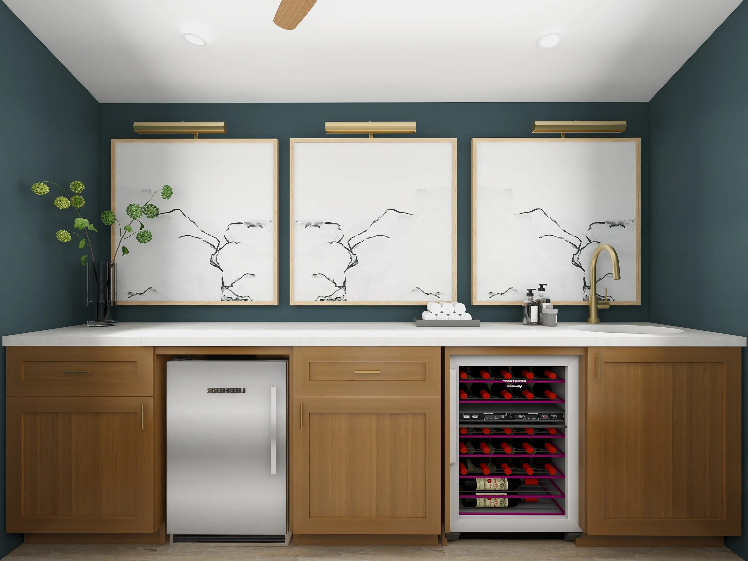 A kitchen with a white counter and wooden cabinetry, featuring a small white refrigerator, wine cooler, and wine rack, with three framed art pieces on the dark teal wall, a plant on the left, and a sink with a gold faucet.