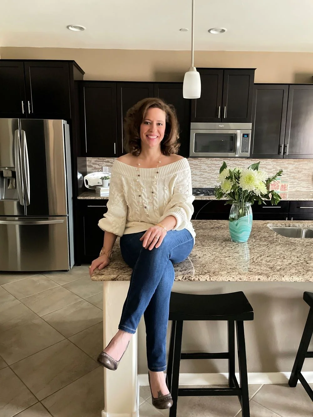 A woman with curly brown hair smiling, sitting on a kitchen counter, wearing a cream sweater with pearl buttons, blue jeans, and brown heels, with a vase of white flowers on the counter behind her.