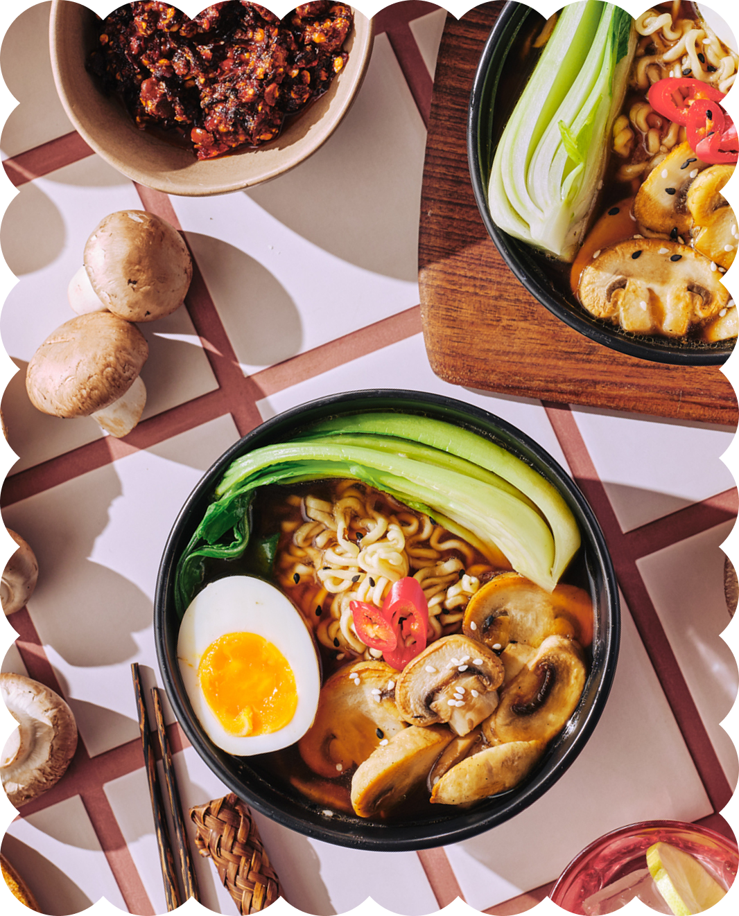 Two bowls of ramen noodles with sliced mushrooms, bok choy, half a soft-boiled egg, and red chili slices, set on a table with mushrooms, a bowl of spicy sauce, and a glass with lemon.