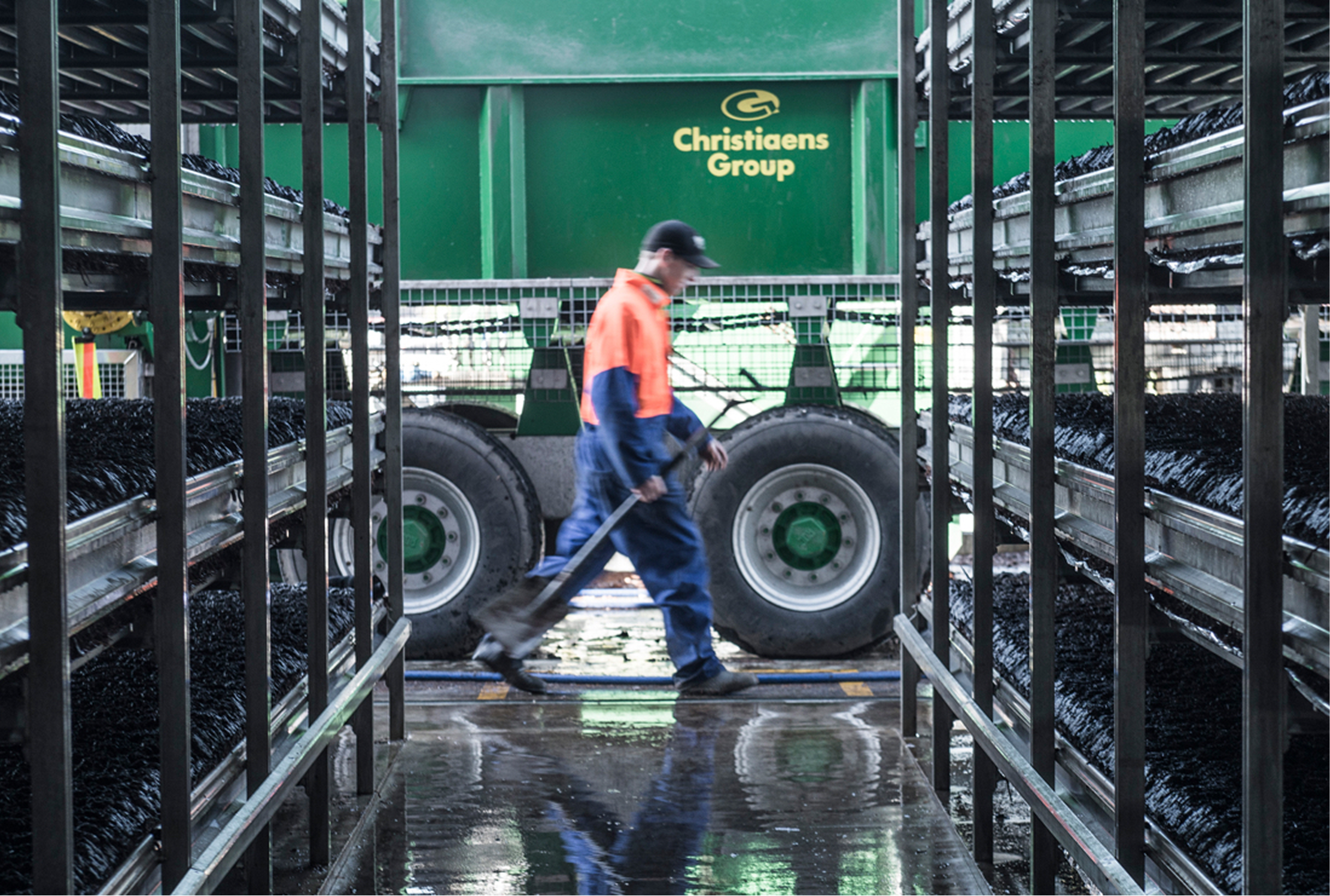 A worker walking through a warehouse aisle with metal shelves filled with black objects, next to a large green truck labeled 'Christiens Group' with only the wheels and part of the truck visible.
