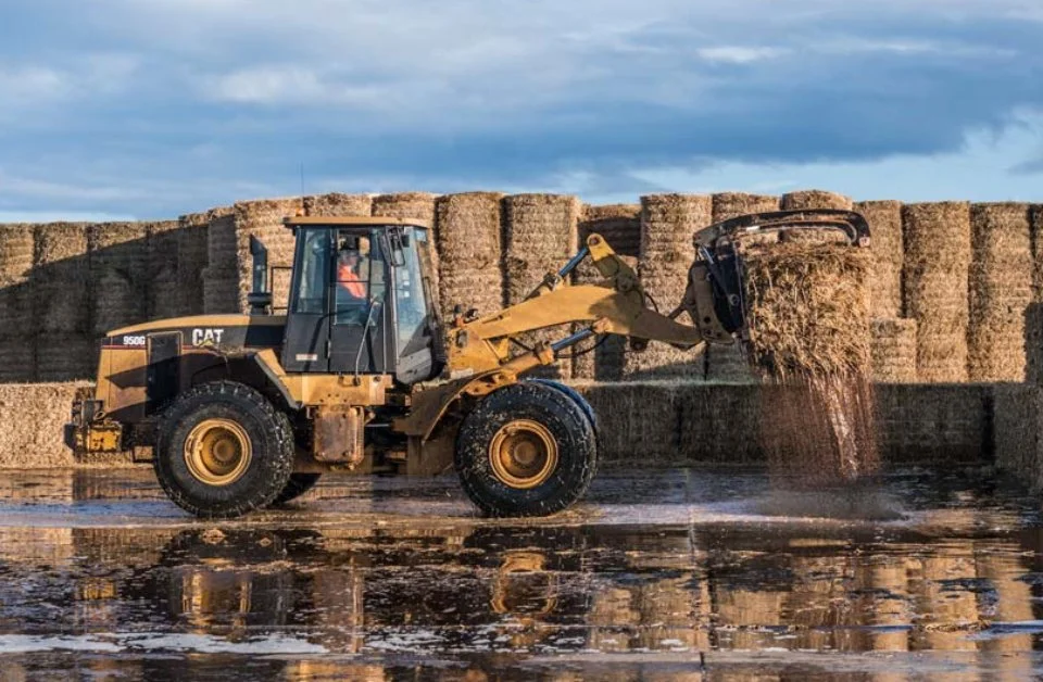 A bulldozer with a loader scoop dumping hay into a large stack of hay bales at a farm, with a haystack in the background and cloudy sky.