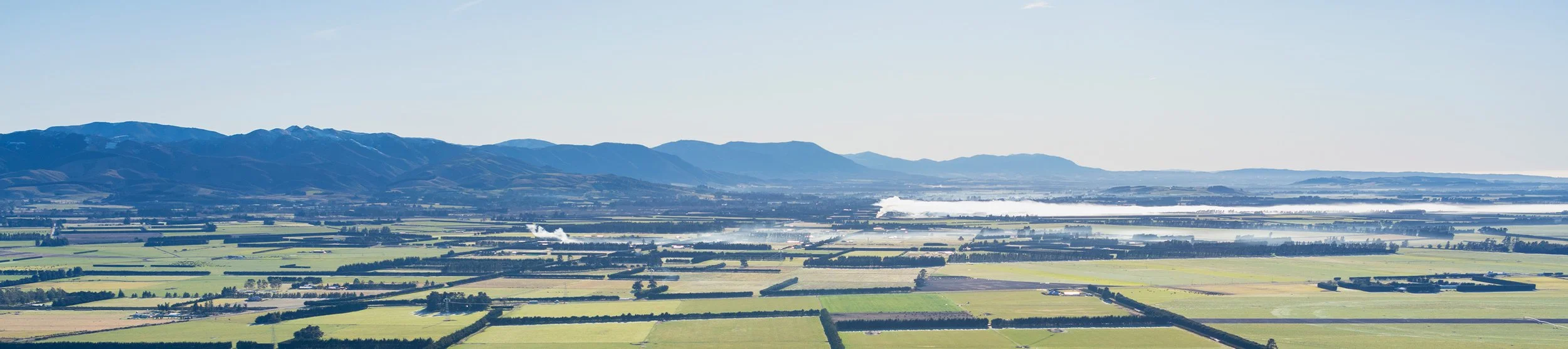 Aerial view of farmland with patchwork fields, a distant mountain range, and smoke rising from some areas on a clear day.