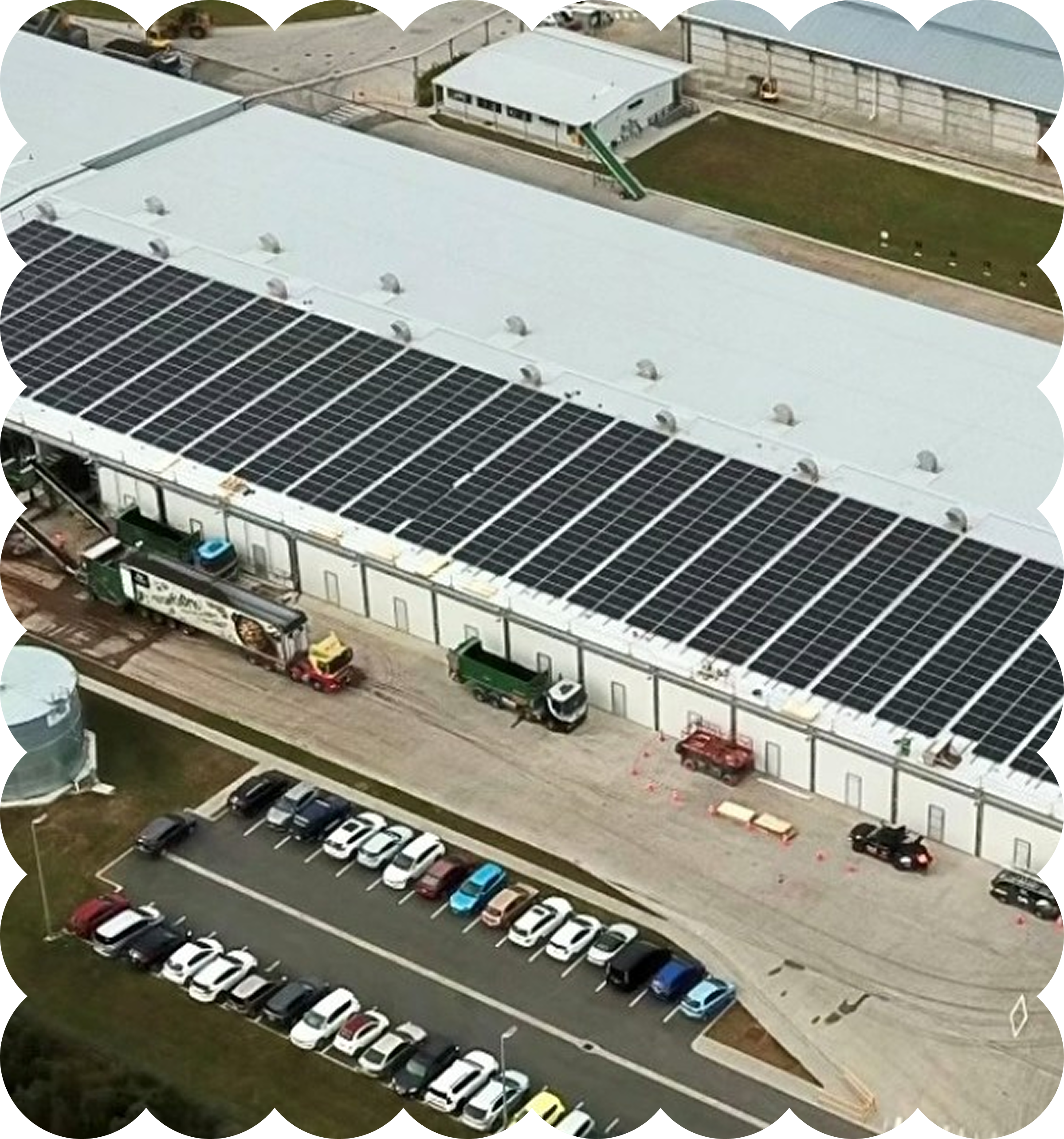Aerial view of a building with solar panels on the roof, a small grass area, a parking lot filled with various colored cars, and a fenced area with a small structure, possibly a warehouse or industrial facility.