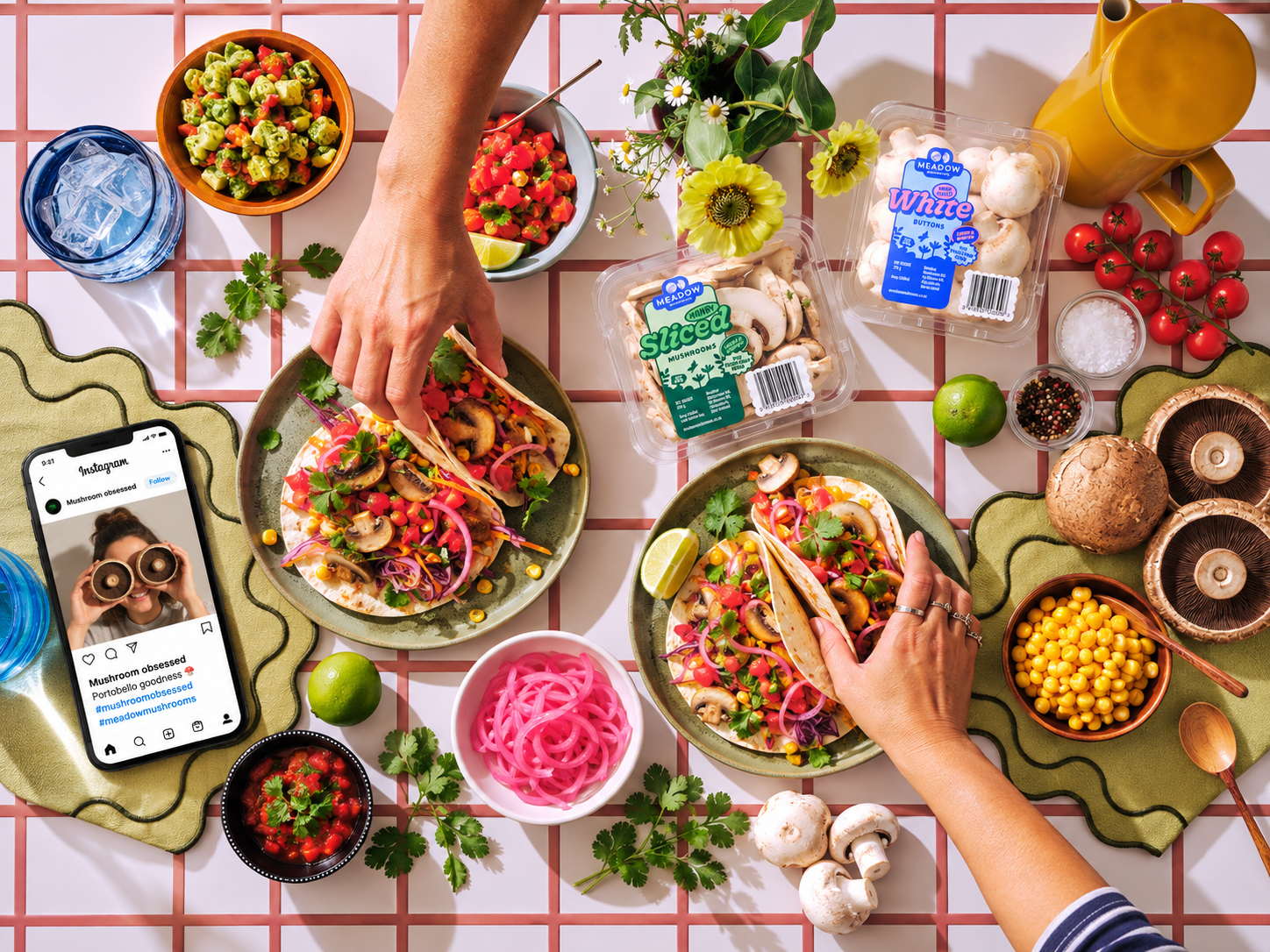 A colorful spread of vegetarian tacos filled with mushrooms, corn, peppers, and cilantro on a checkered table. Surrounding the tacos are fresh ingredients like cherry tomatoes, mushrooms, lime, and onions, alongside spices, a wedge of lime, a bowl of pink pickled onions, a box of sliced mushrooms, and a bottle of hot sauce. A smartphone showing an Instagram post about mushrooms is also on the table.