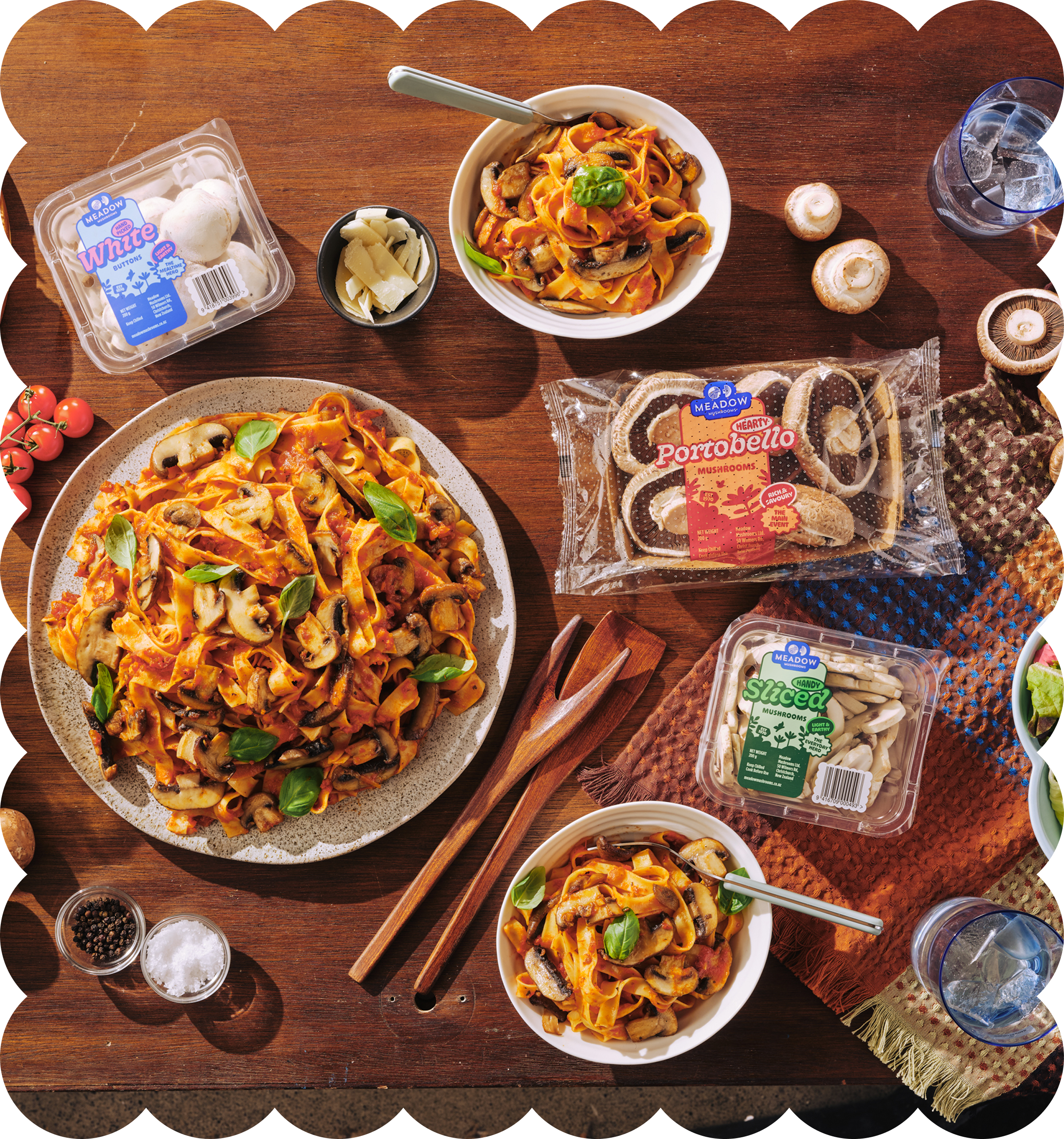 Top-down view of a wooden table with plates of mushroom pasta, a small bowl of cheese, a bowl of peppercorns, and slices of portobello mushrooms, along with a glass of iced water and a package of sliced mushrooms.
