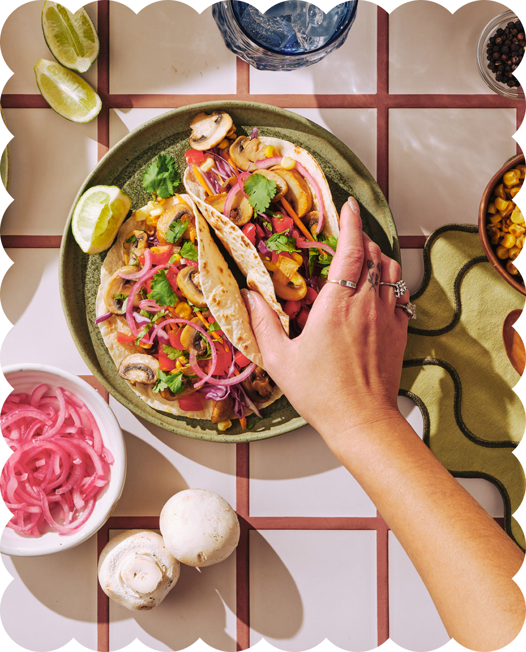 Person placing a taco filled with mushrooms, corn, onions, and cilantro onto a plate, surrounded by lime wedges, sliced onions, mushrooms, water, and other bowls of ingredients on a tiled table.