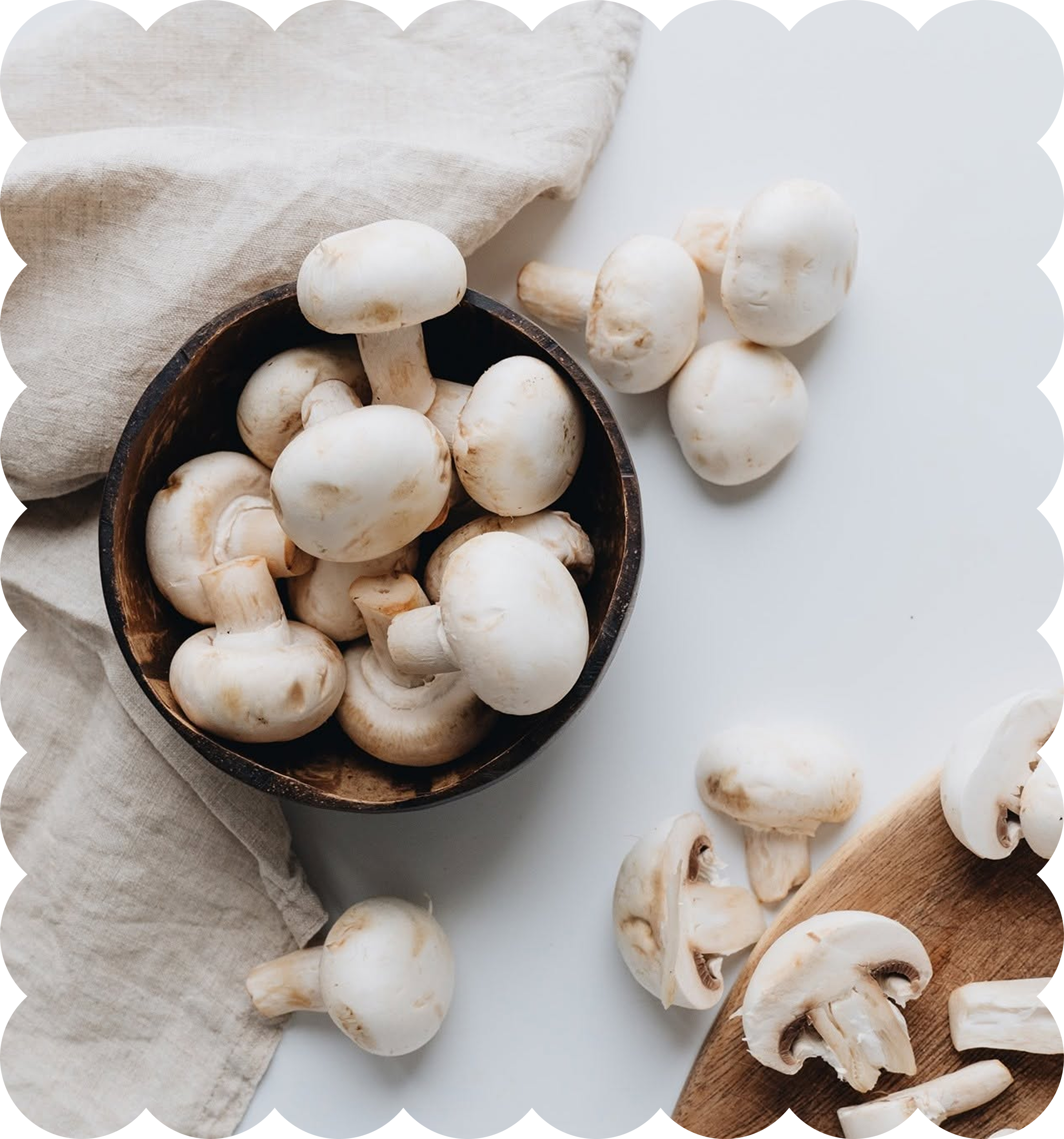 Fresh white mushrooms on a bowl, a pile on a wooden spoon, and some sliced on a cutting board with a beige cloth nearby.
