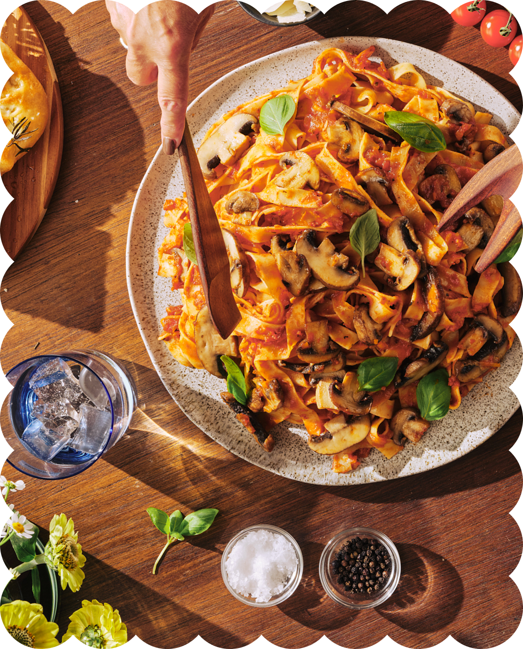 A plate of cooked fettuccine pasta with mushroom, tomato sauce, and fresh basil leaves on a wooden table, with salt, black pepper, and a glass of ice water nearby.