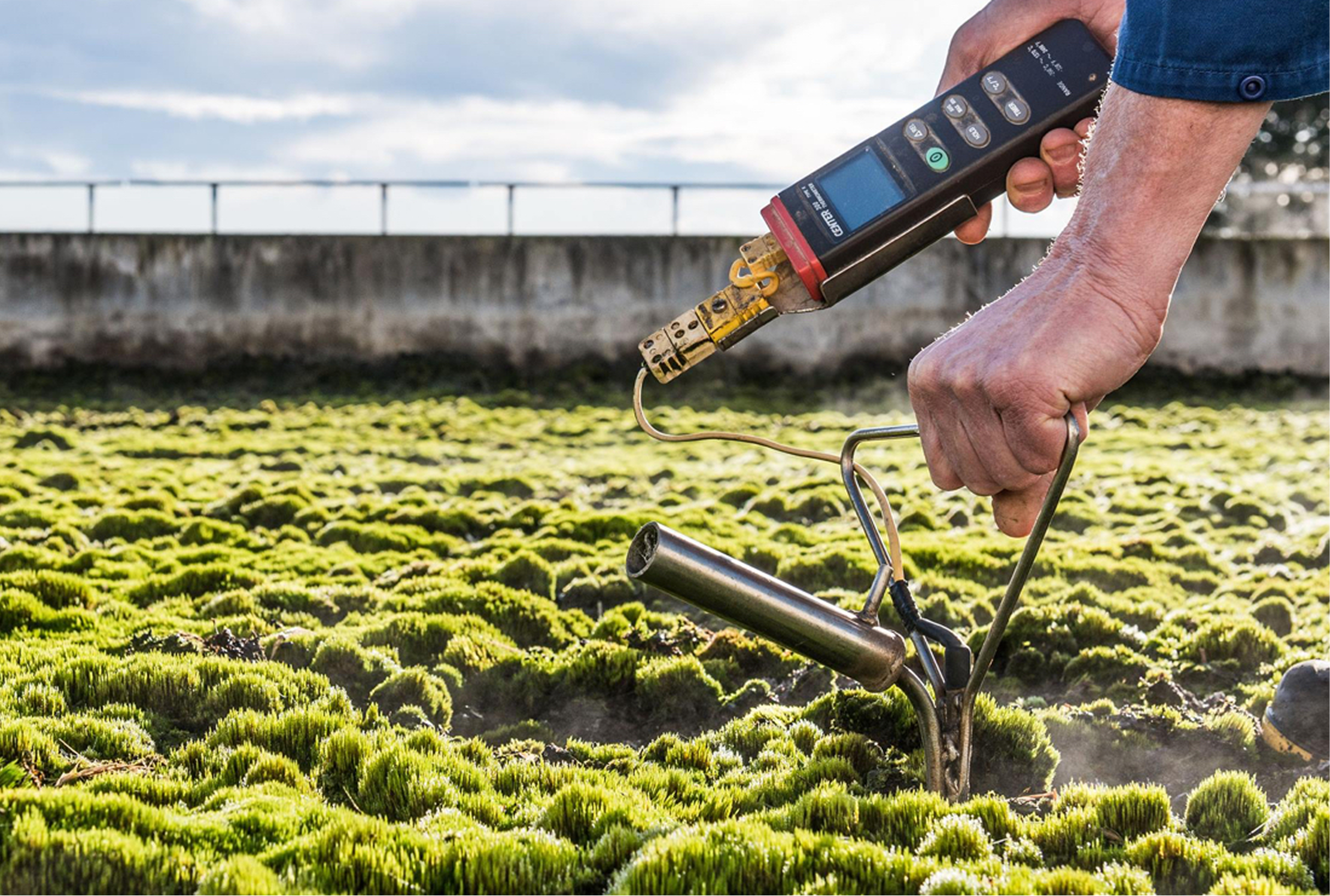 Farmer using soil testing device in a field of green plants with a bridge and cloudy sky in the background.